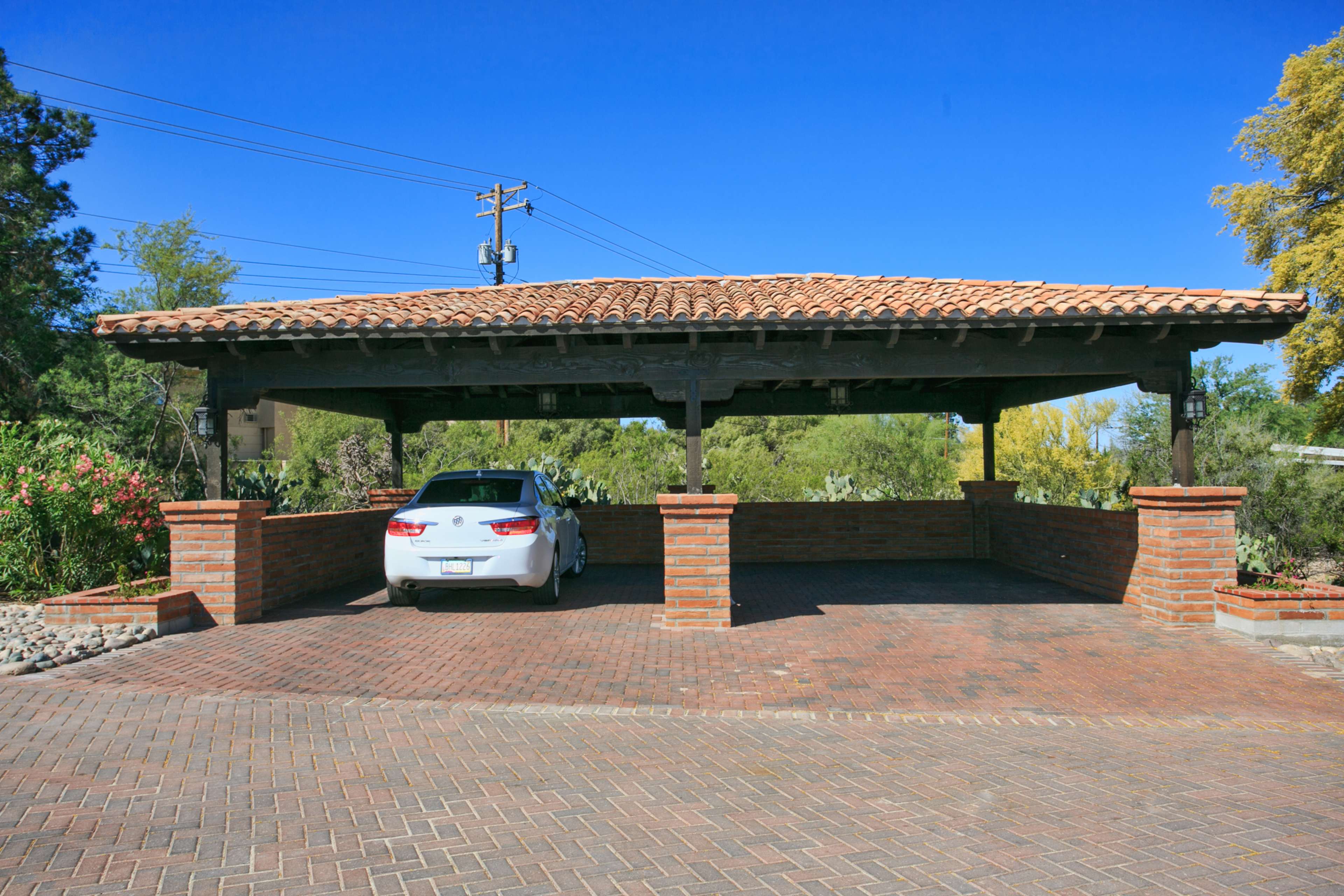 A white car is parked under a tiled roof carport constructed with brick columns and surrounded by landscaped greenery.