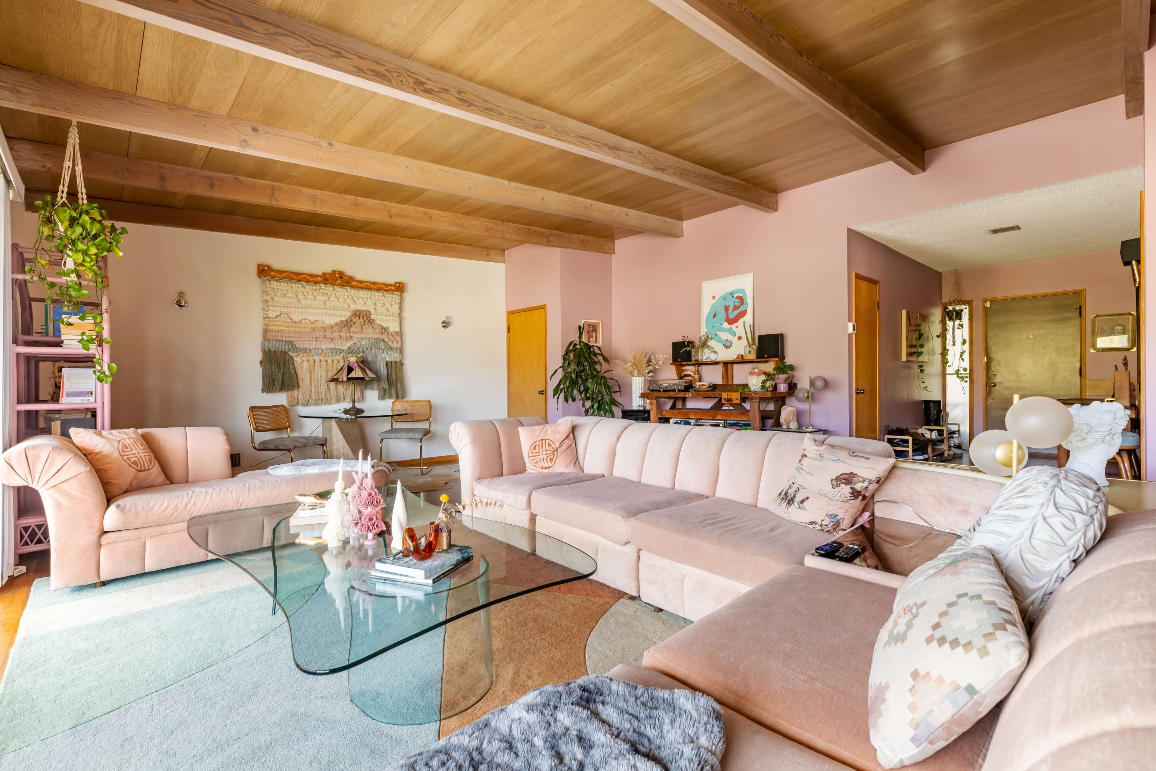 A spacious living room features a large, curved pink sectional sofa, a glass coffee table, and a dining area in the background.