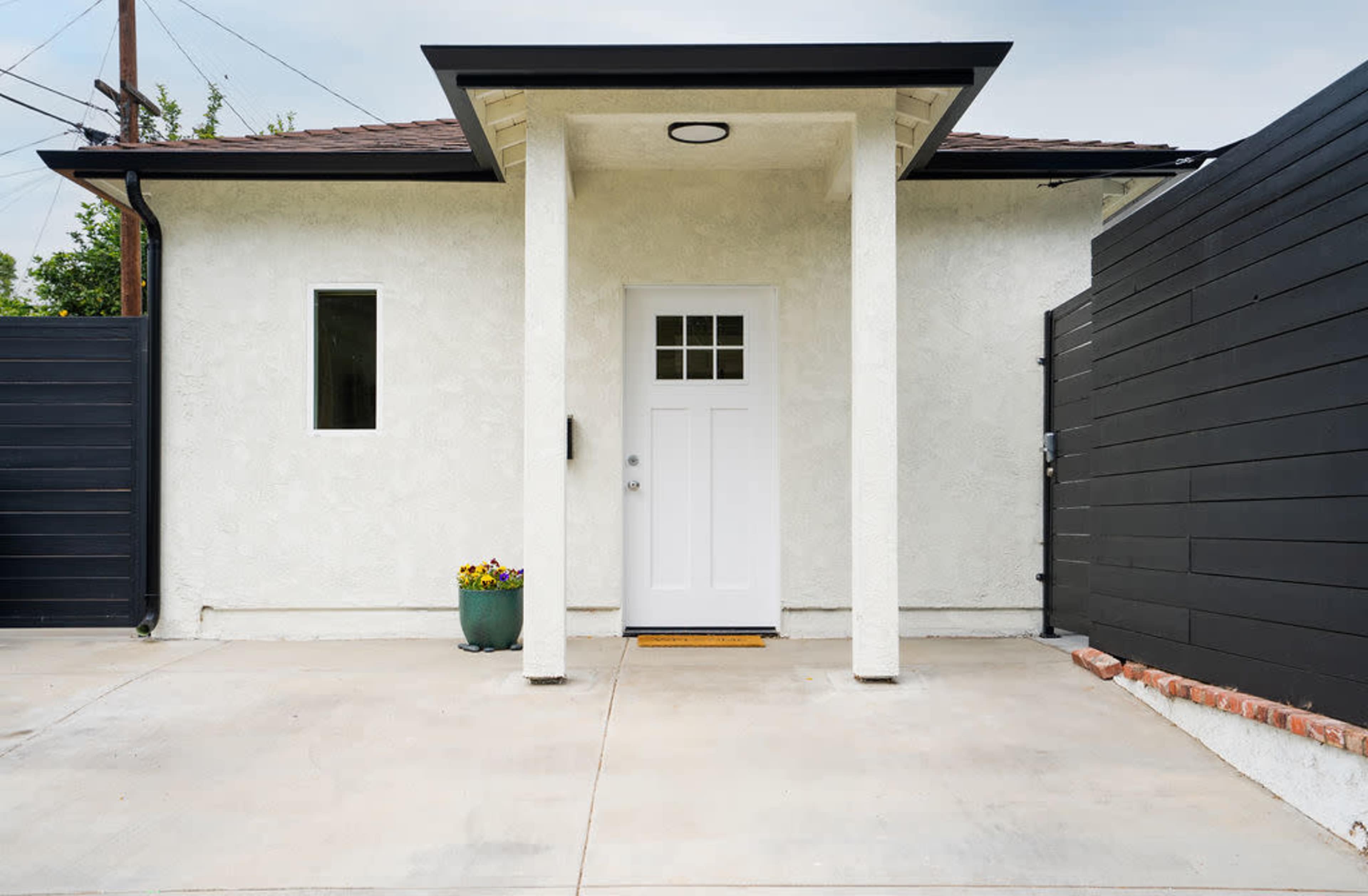 A modern entrance to a house with a white door, a porch with a roof, and dark wooden fencing on either side.