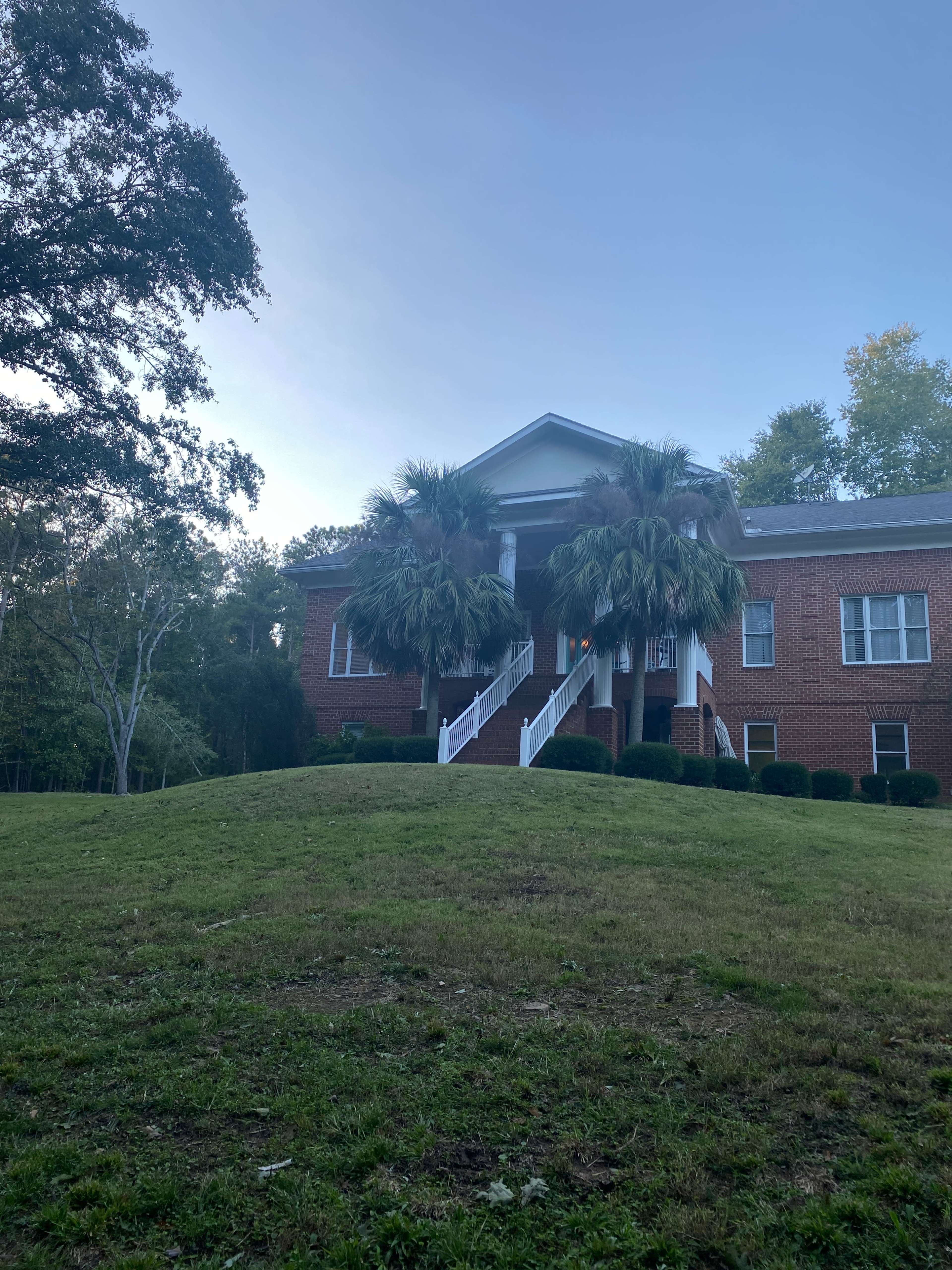 A brick house with a white staircase and palm trees is situated on a grassy hill.