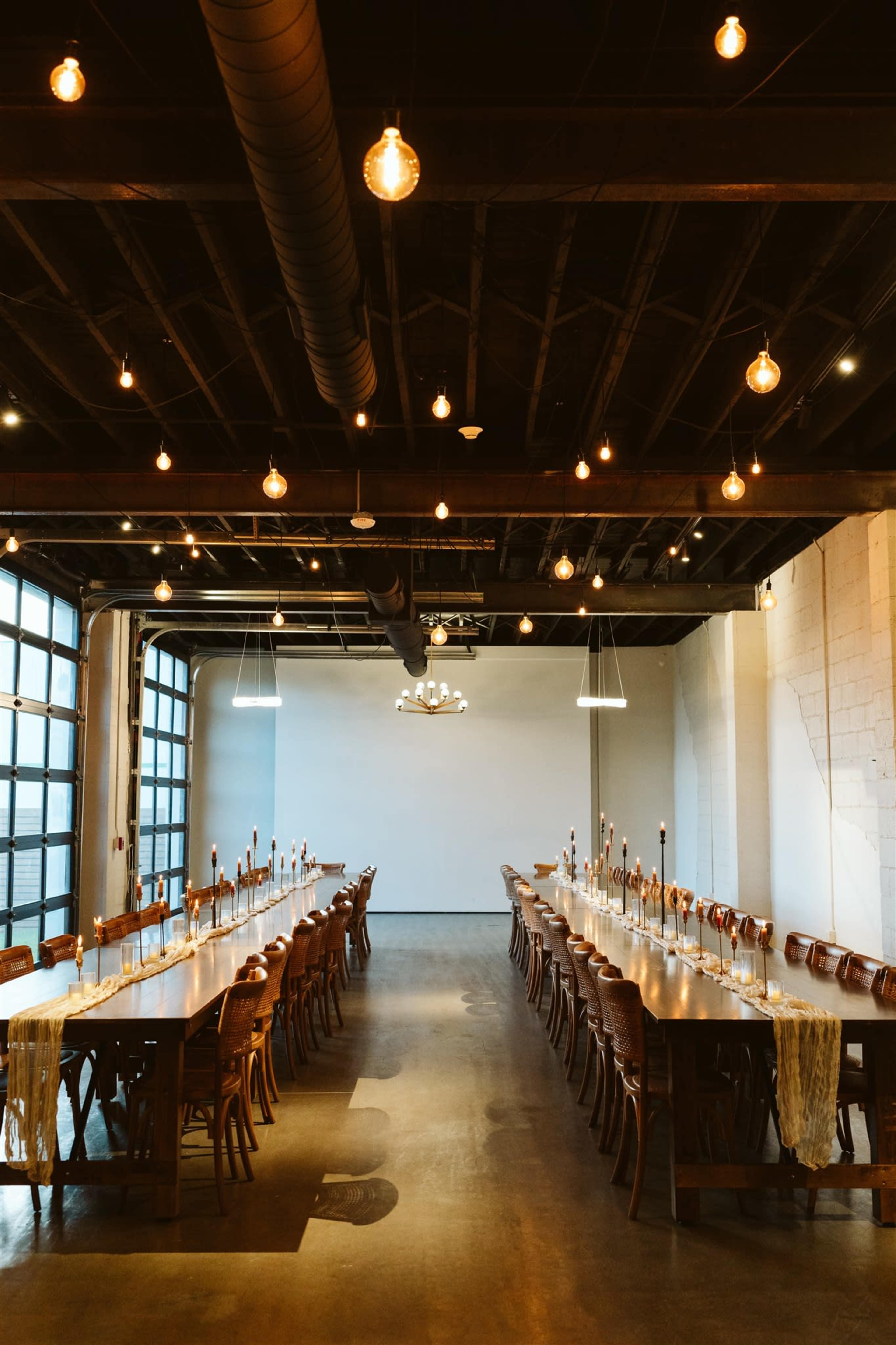 A long wooden table with chairs is set in a spacious room with industrial-style lighting and large windows.