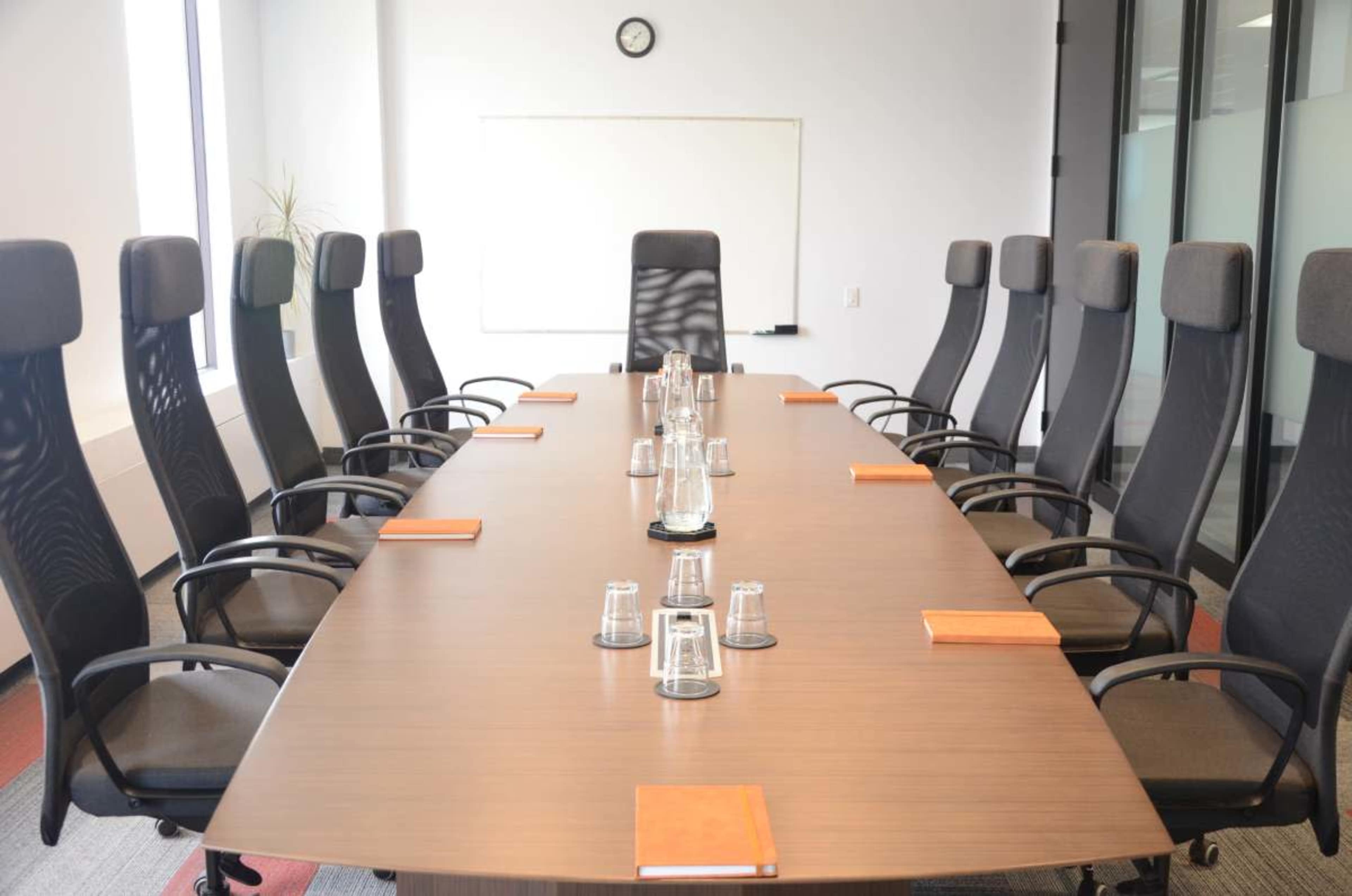A large conference room features a long brown table surrounded by ergonomic black chairs, with notebooks and glassware placed along its length.