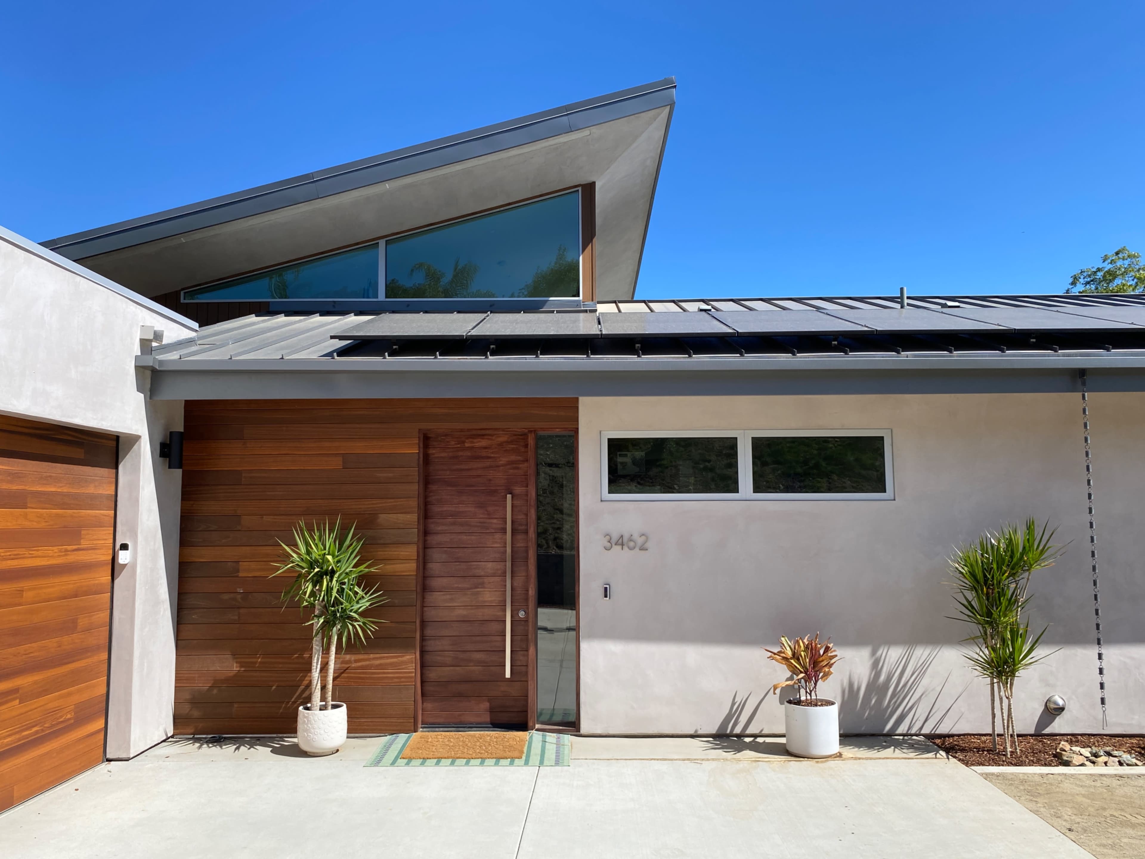 The image shows a modern house with a wooden door, concrete walls, and solar panels on the roof, surrounded by landscaped plants.