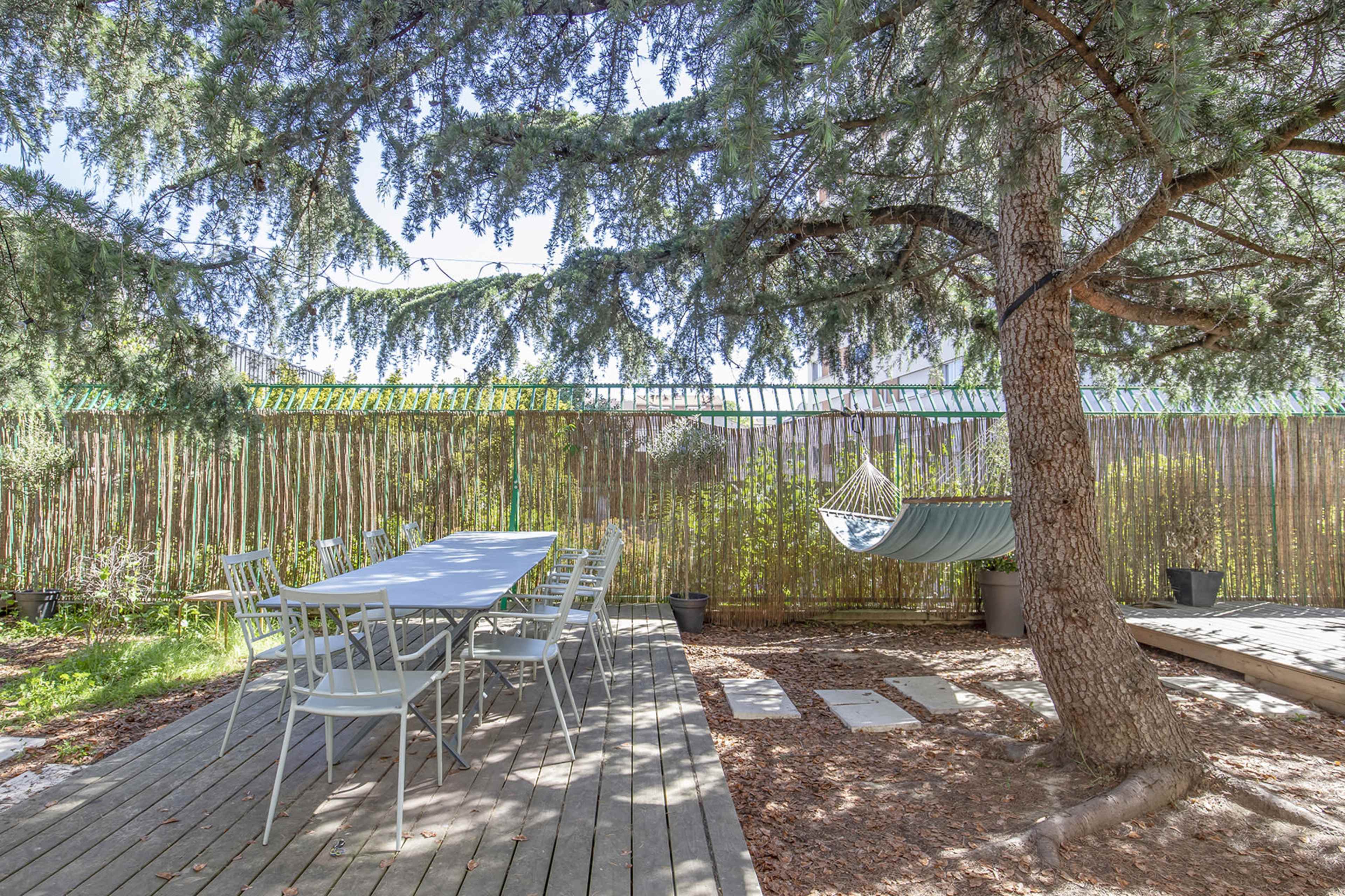 A rectangular dining table with six chairs is set on a wooden deck beneath a tree, alongside a hammock hanging between two posts in a shaded outdoor area.