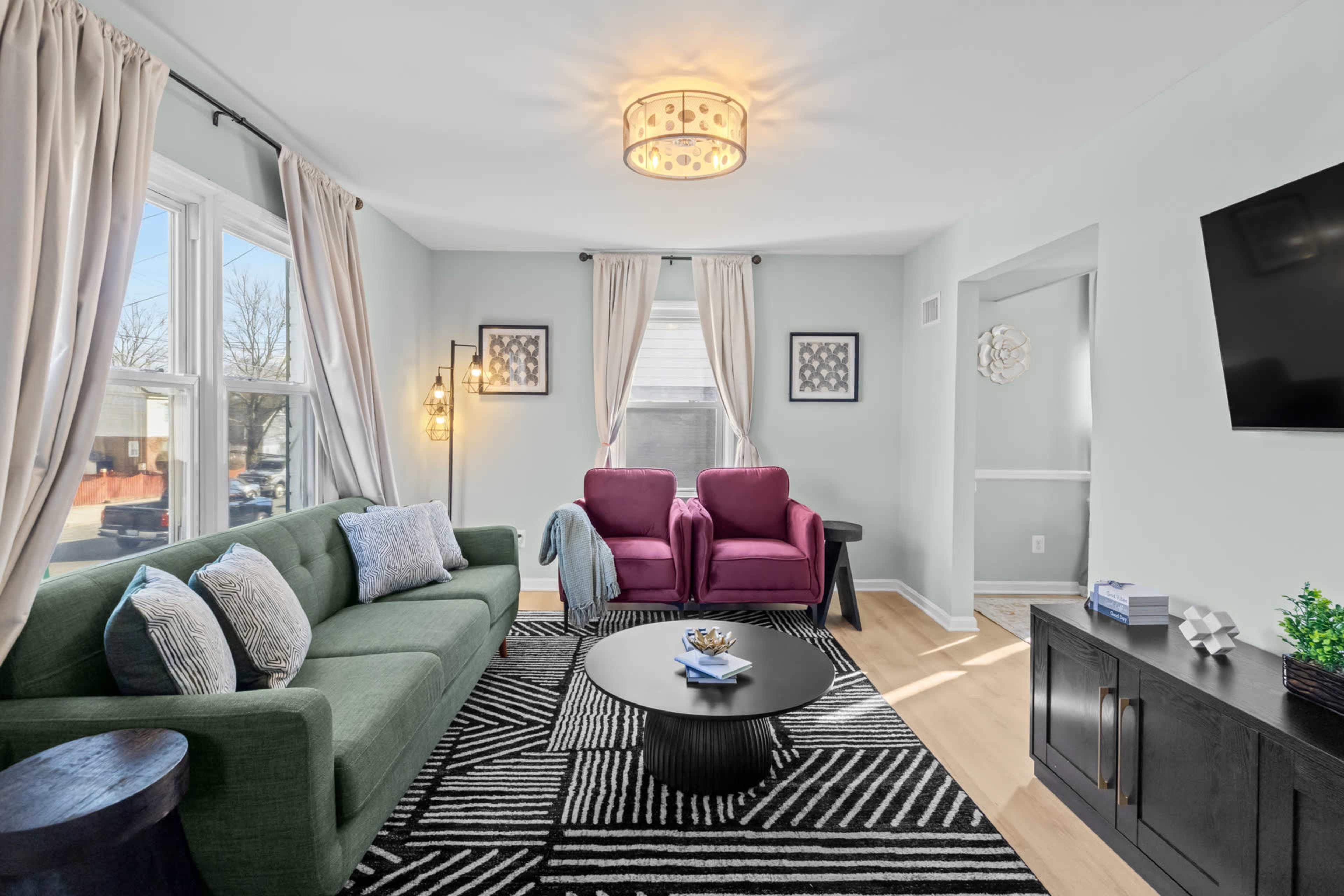 The image shows a modern living room featuring a green sofa, two purple armchairs, a round coffee table, and a television mounted on a wall.