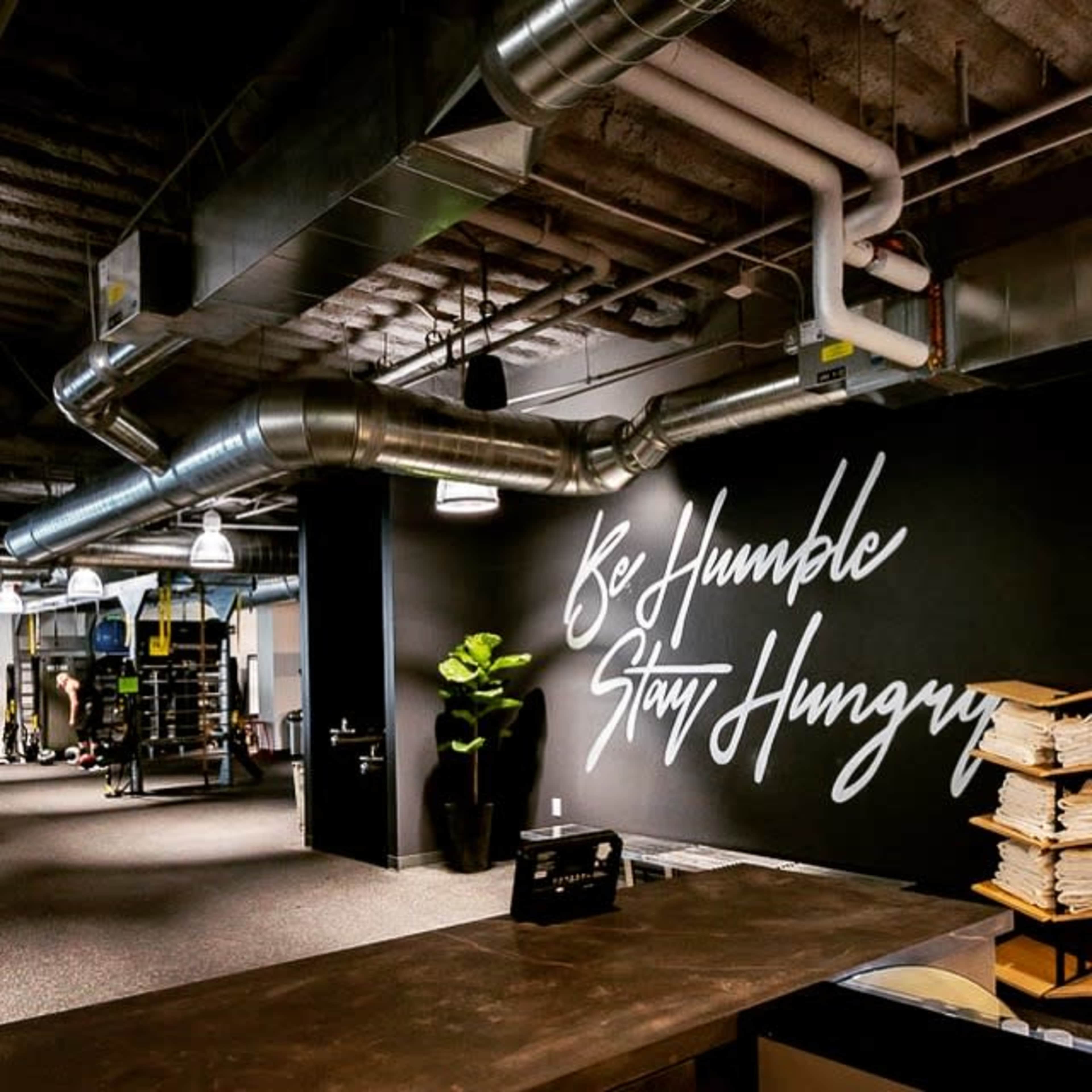 The image shows a gym interior featuring a wall with the phrase "Be Humble Stay Hungry" in stylish lettering, along with various workout equipment and a reception area.