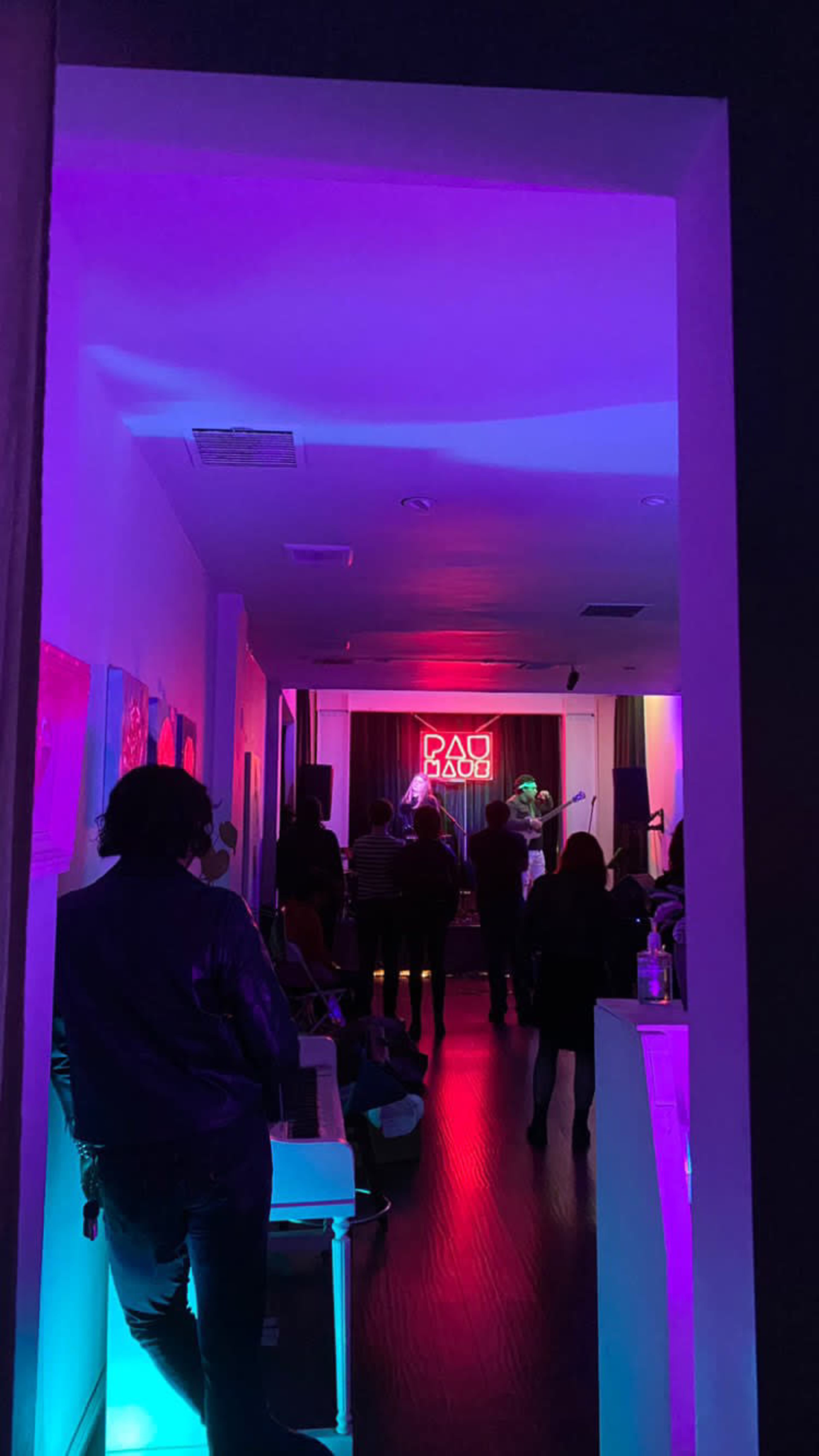 A small music venue with a stage in the background illuminated by purple and pink lights, filled with an audience watching a performance.