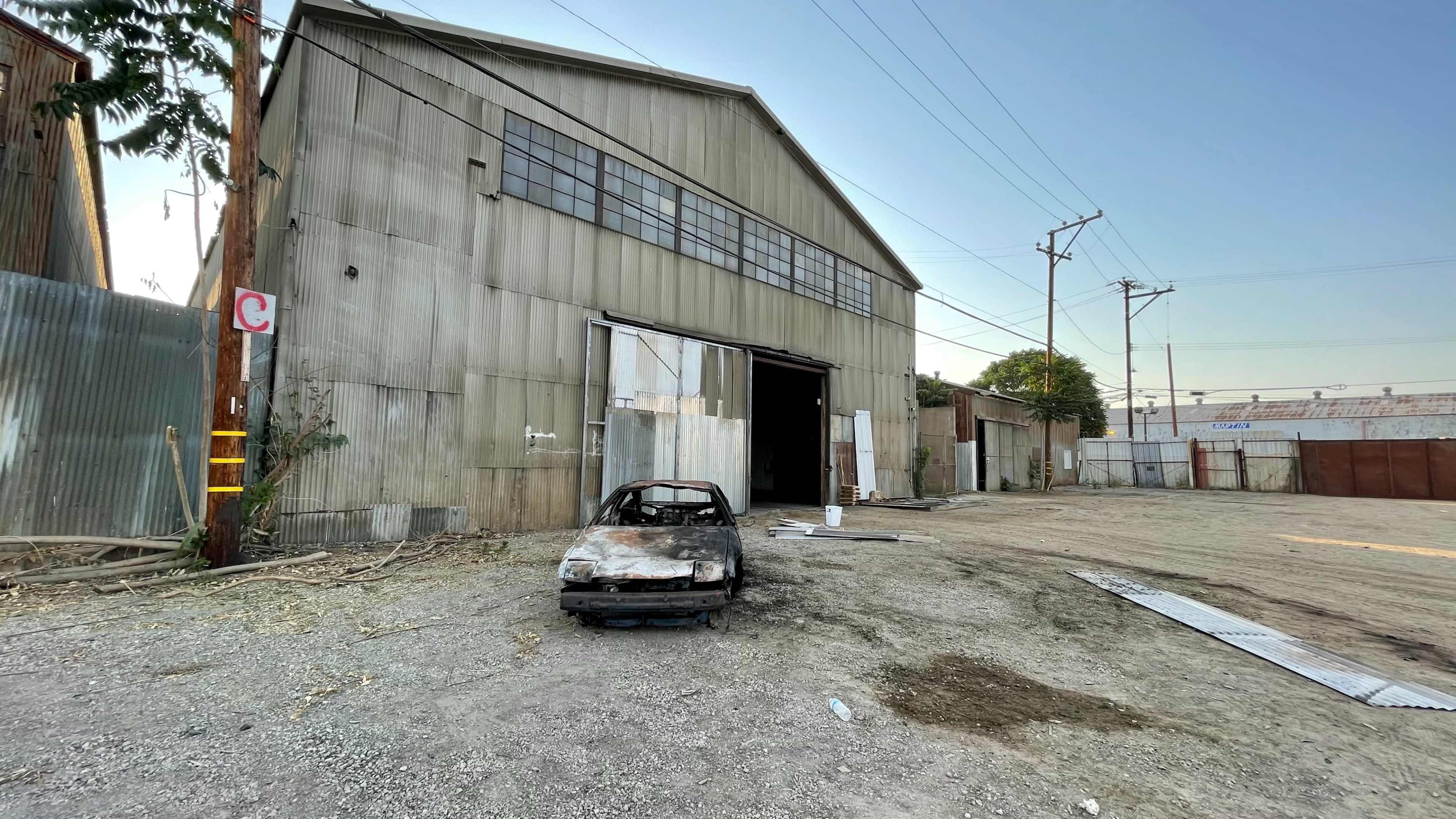 An old, burned-out car sits in front of a large, corrugated metal warehouse in a gravel lot.