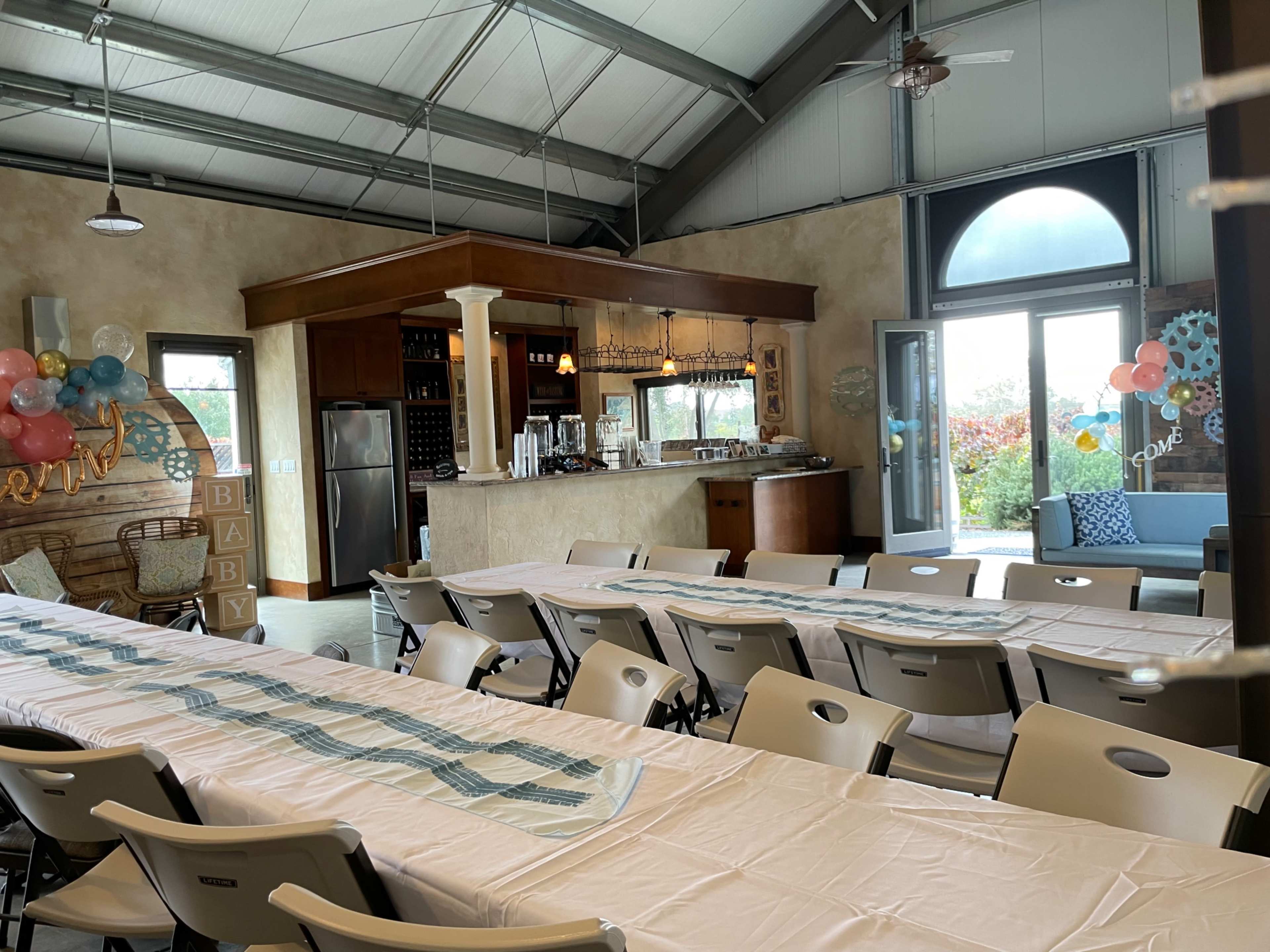 An indoor party venue features a long table set for guests, with a kitchen area in the background and decorations displayed along the walls.