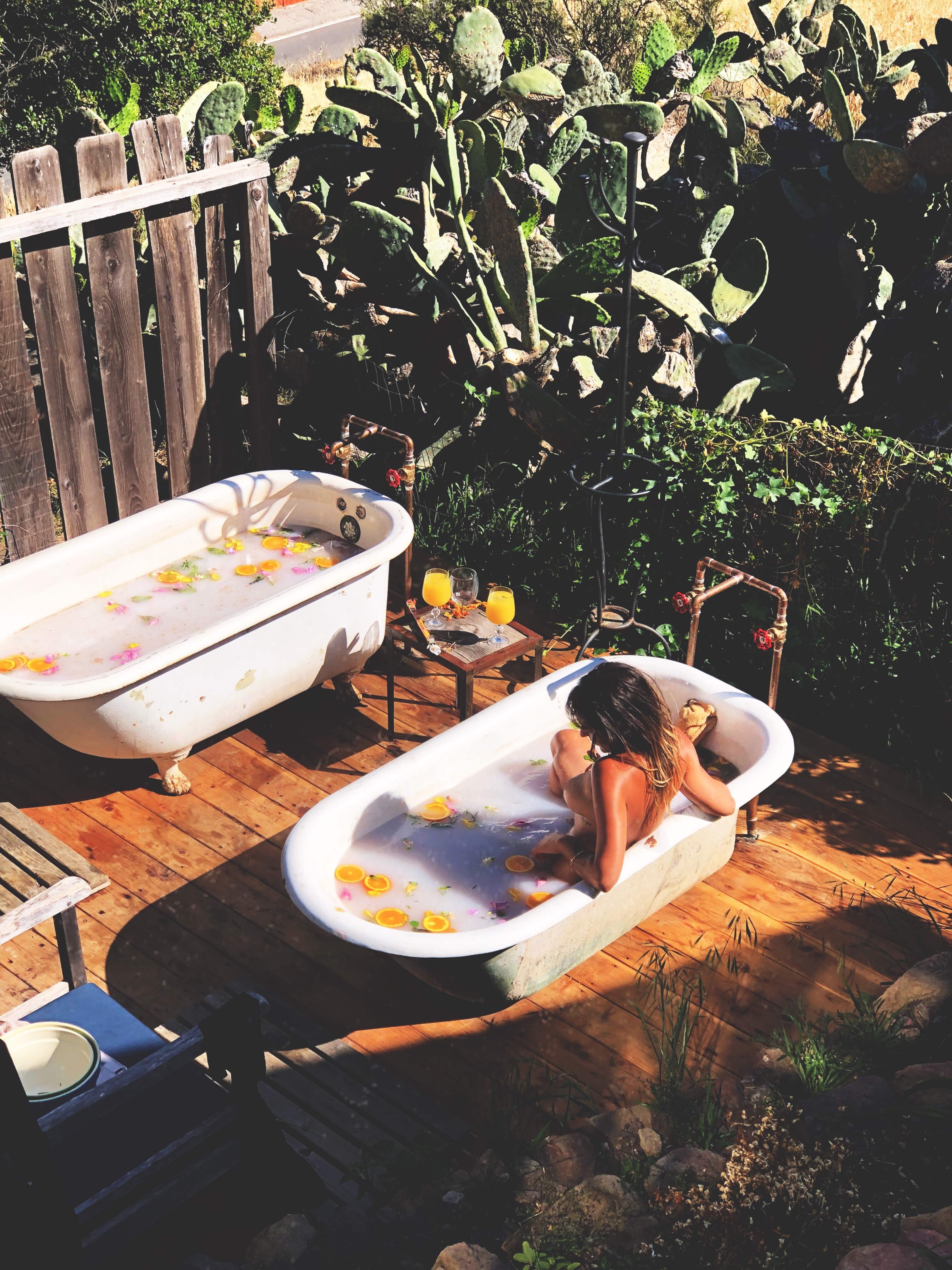 Two vintage bathtubs filled with water and flower petals sit on a wooden deck surrounded by cacti, while a person relaxes in one of the tubs.