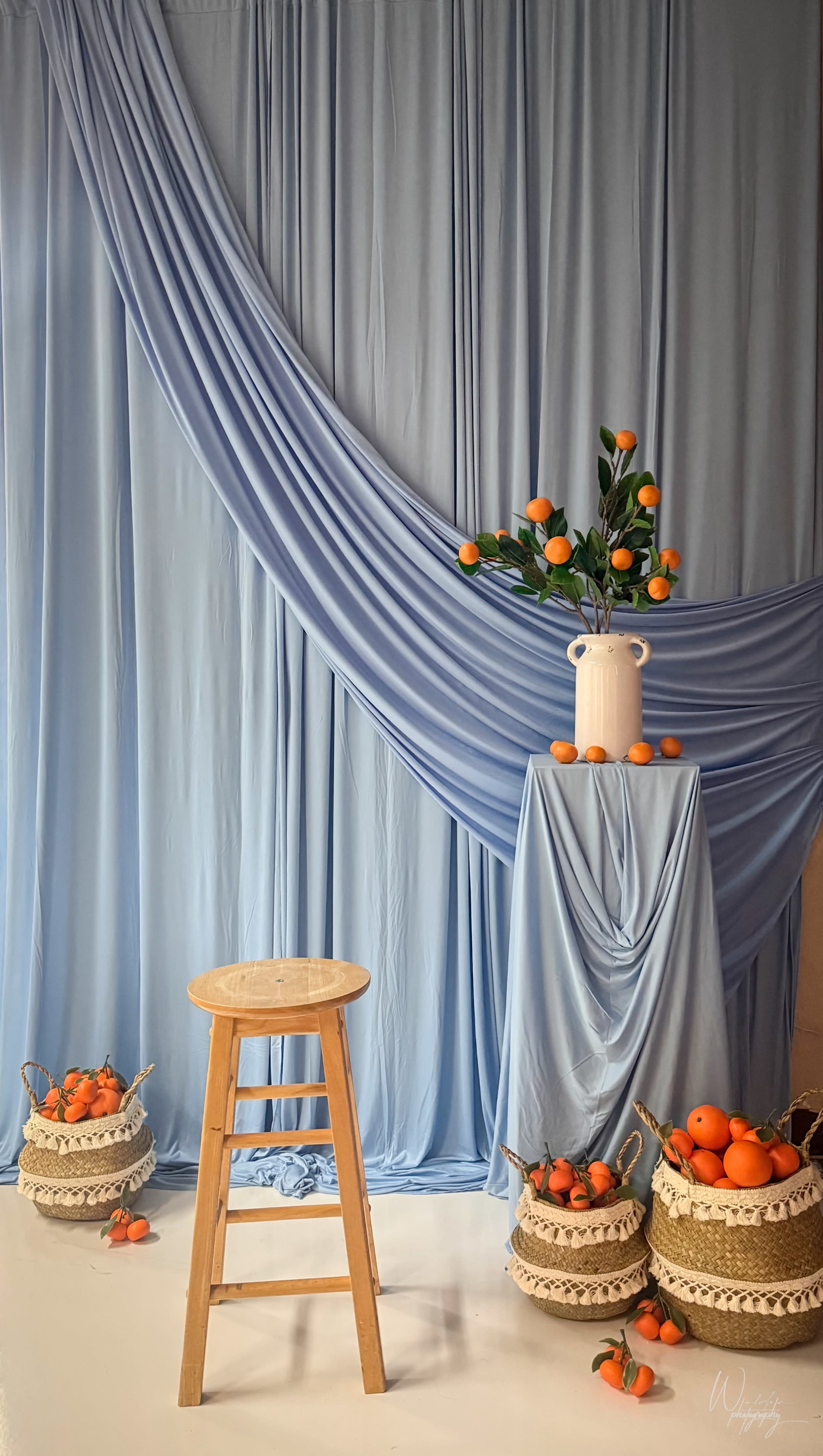 The image features a light blue draped backdrop with a wooden stool, a table decorated with a white vase holding greenery and orange fruits, and two woven baskets filled with oranges.