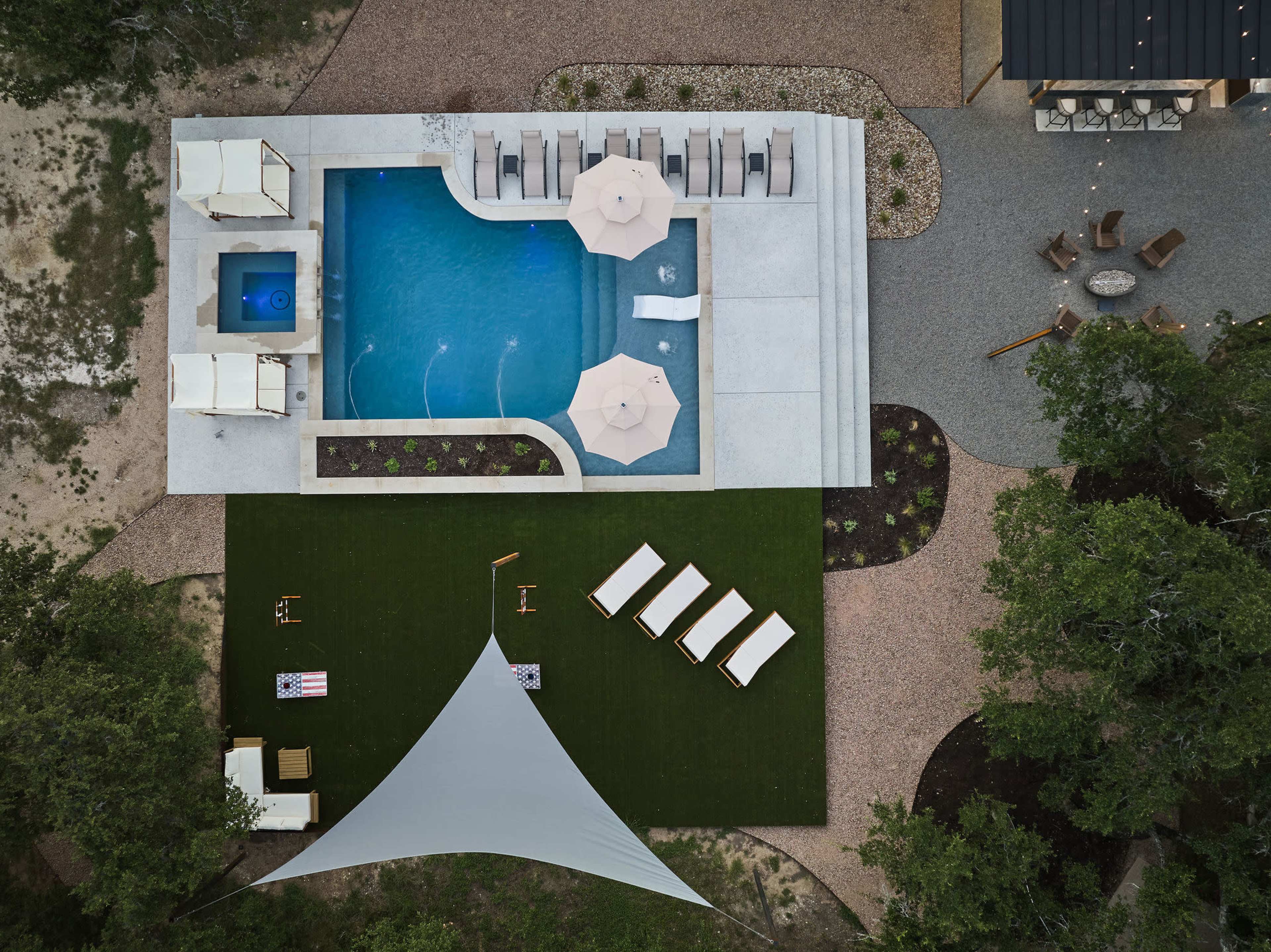 The image shows an aerial view of a rectangular pool surrounded by lounge chairs, umbrellas, and a green lawn area, with a tent structure nearby and a fire pit in the background.