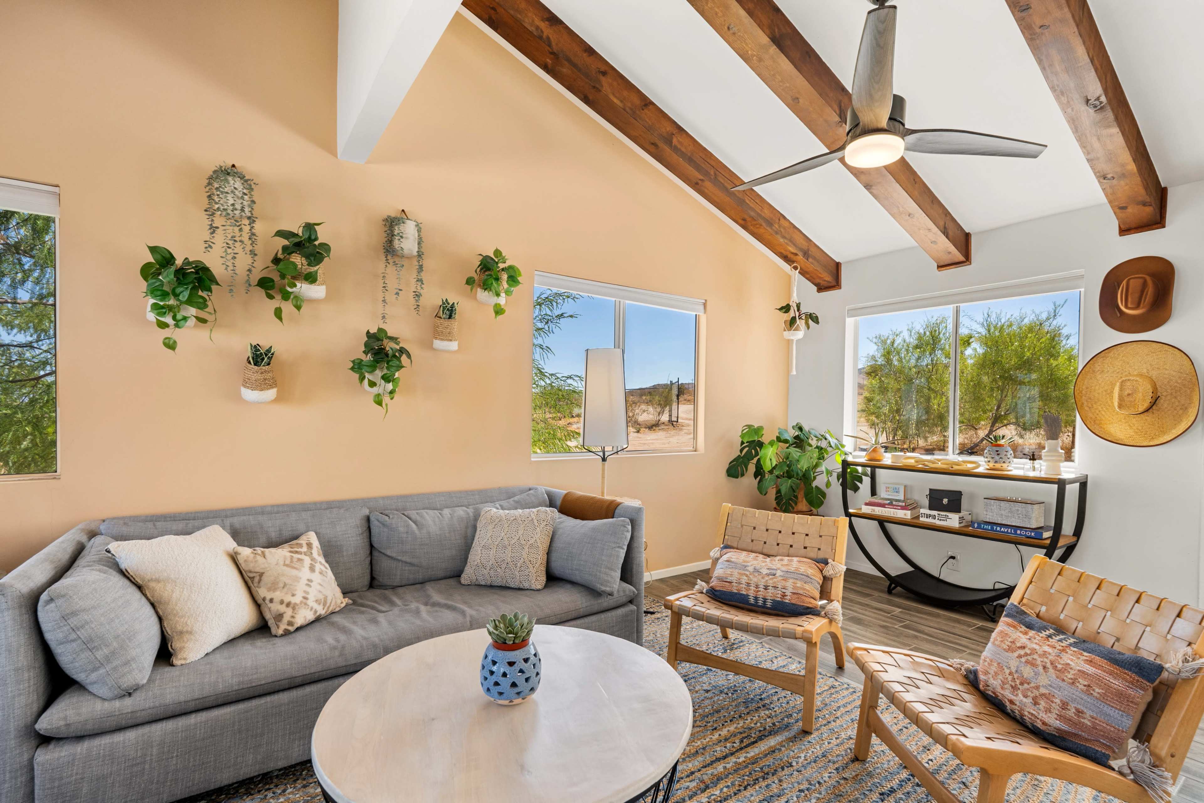 A cozy living room features a gray sectional sofa, two woven chairs, a round coffee table, and various plants arranged on the walls and furniture.