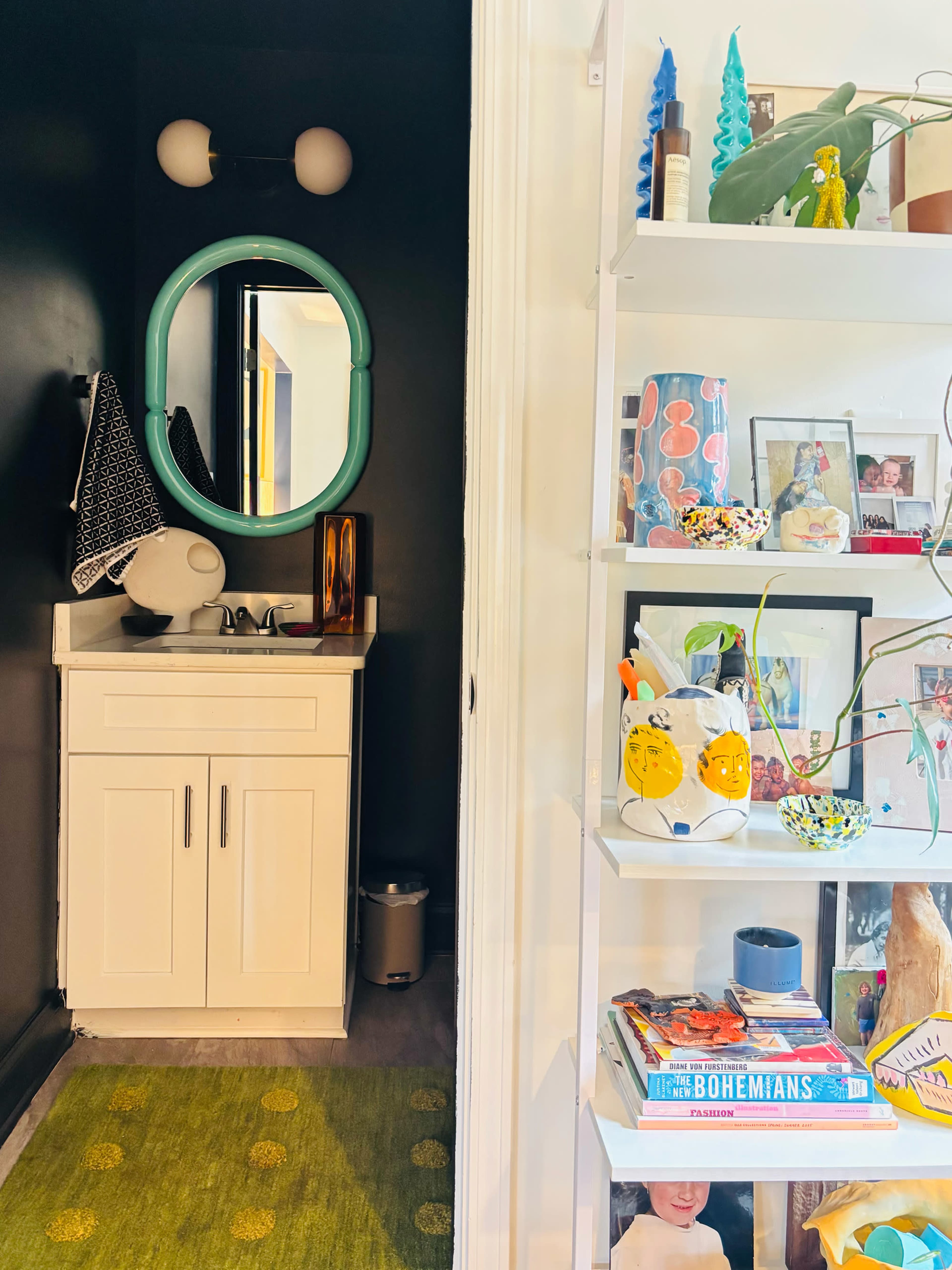 A narrow hallway features a white vanity with a sink and a green-framed mirror on one side, while a bookshelf filled with colorful items and photographs sits on the opposite side.