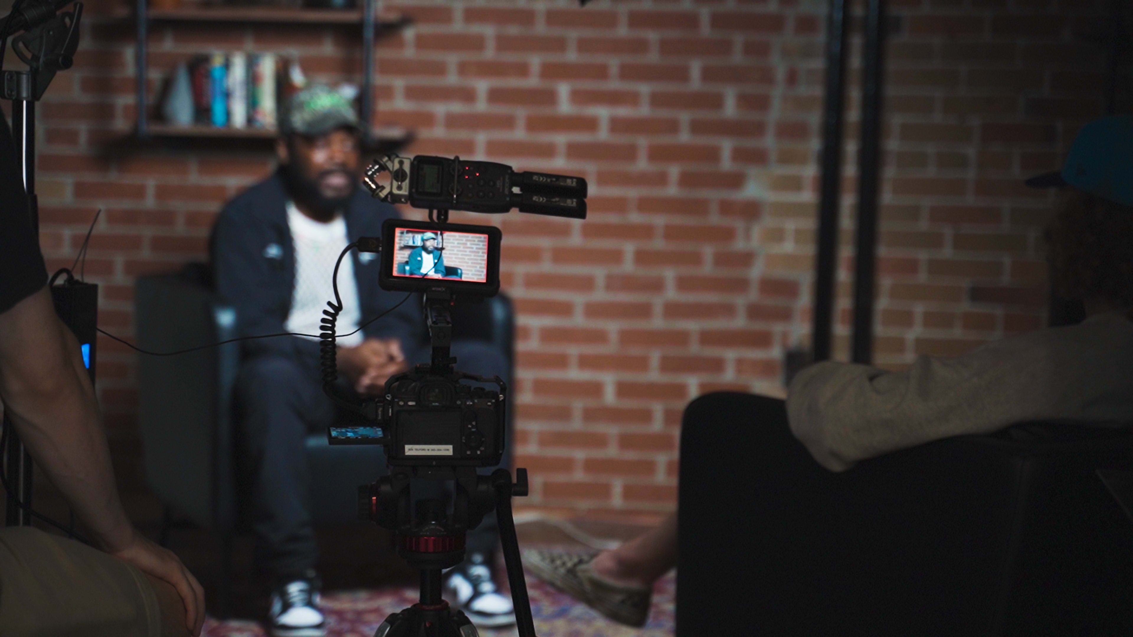 A video interview is being filmed with a seated subject in front of a brick wall, while a camera adjusts to capture the scene.