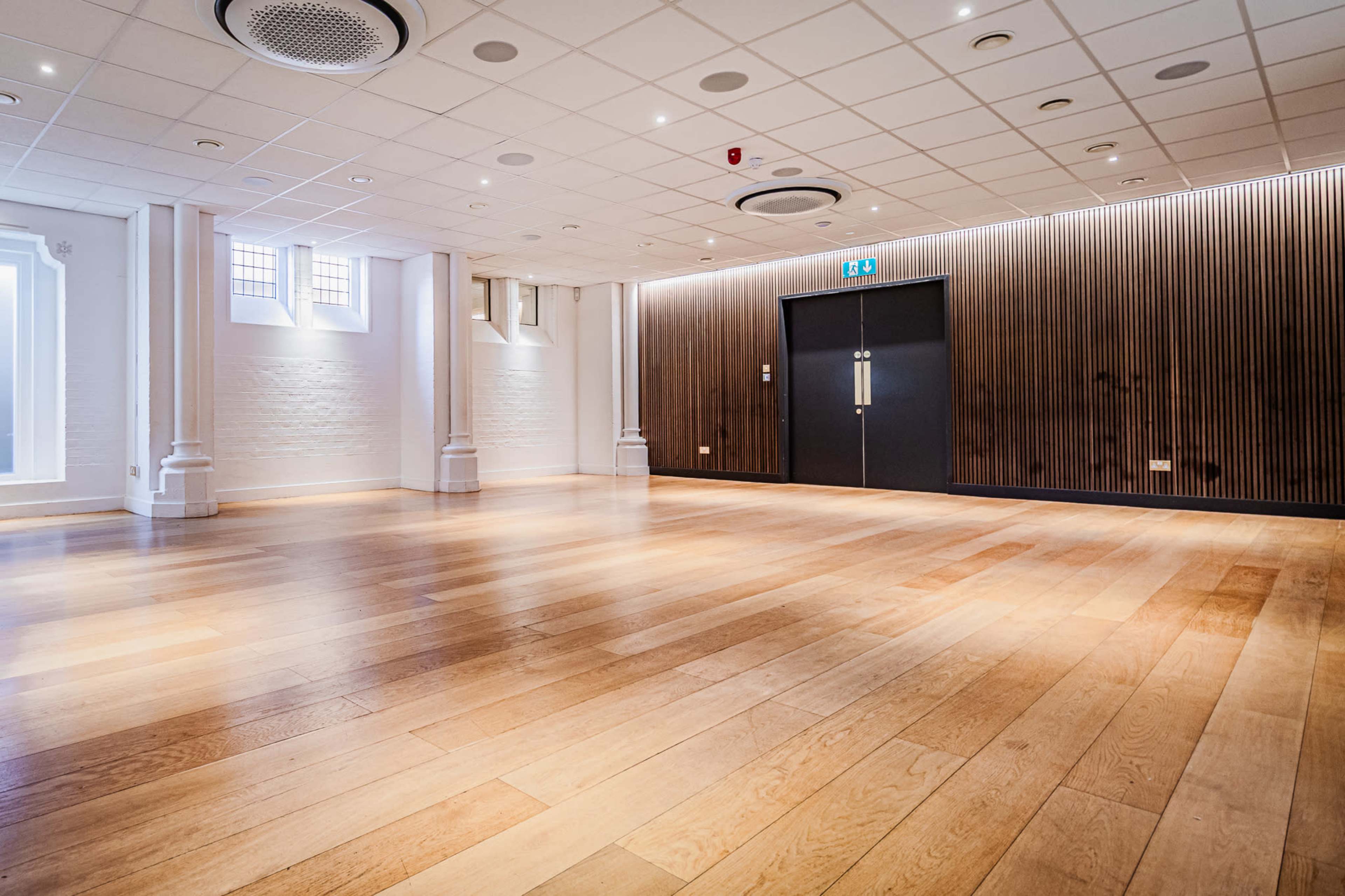 The image shows an empty, well-lit room with polished wooden flooring and a dark wooden feature wall.