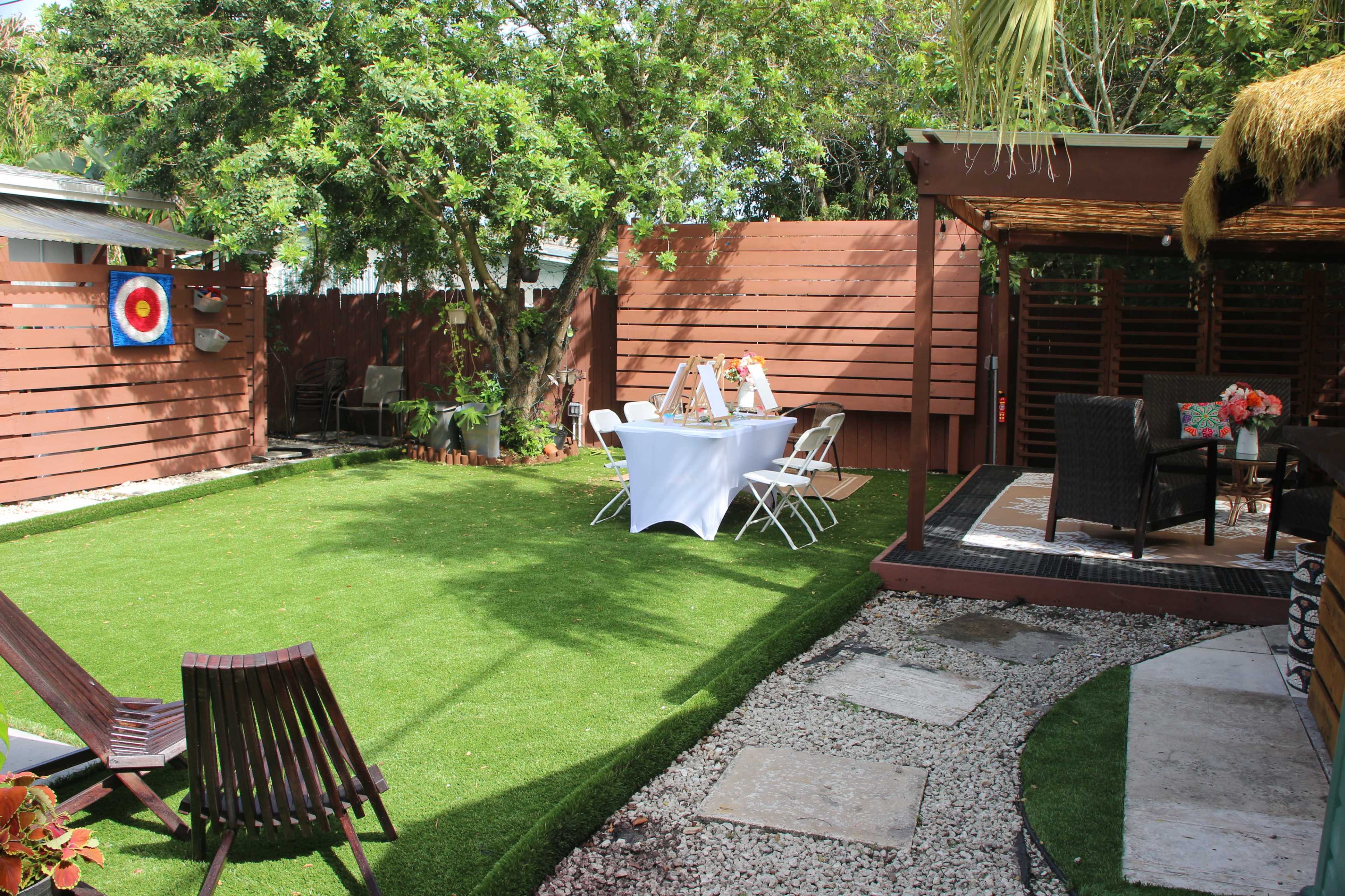 The image shows a well-maintained backyard with artificial grass, a seating area under a wooden structure, and a table set for an outdoor event.
