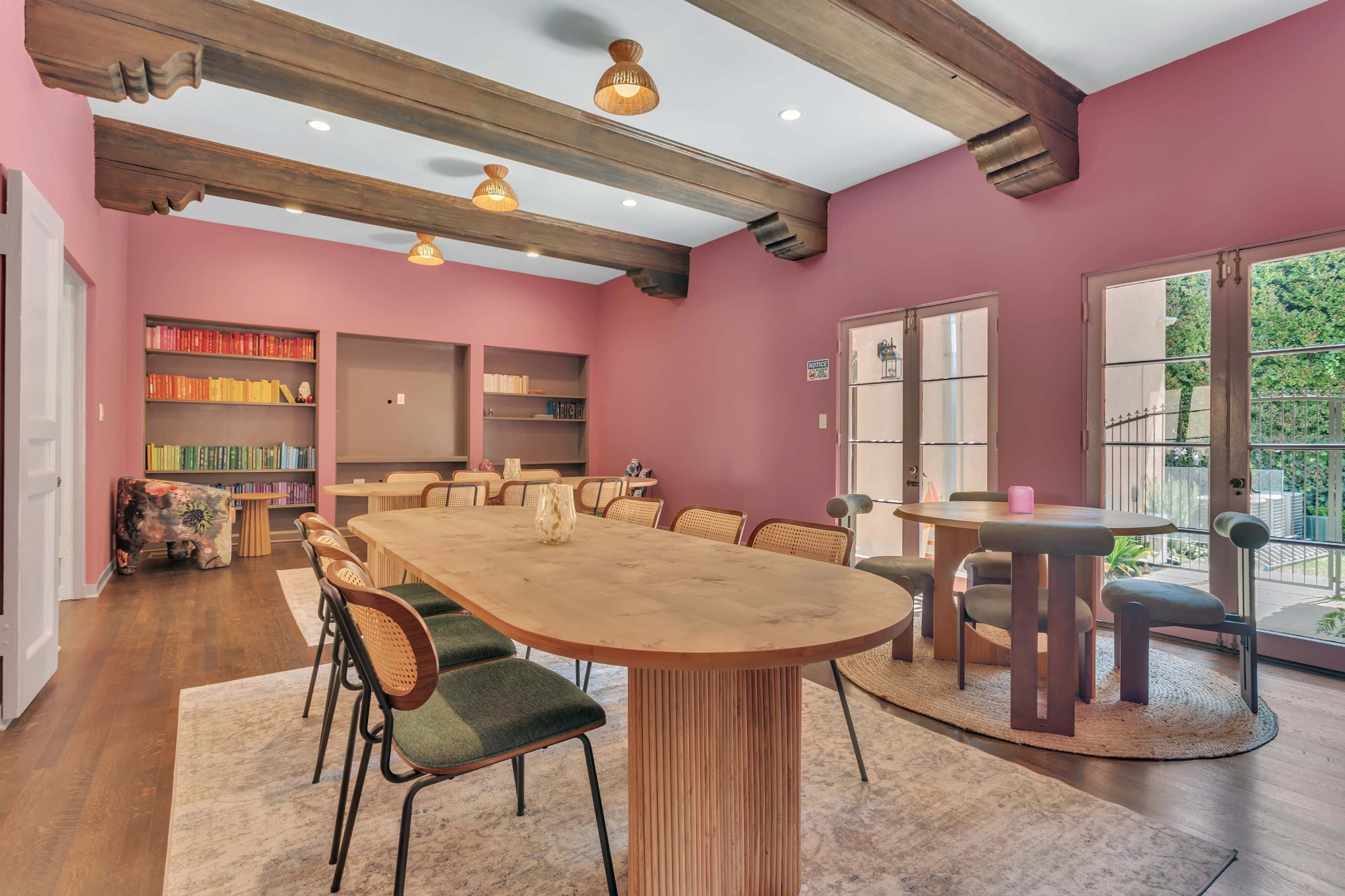 A spacious room features a central oval table surrounded by chairs, with a pink accent wall, wooden beams, and bookshelves filled with colorful books.