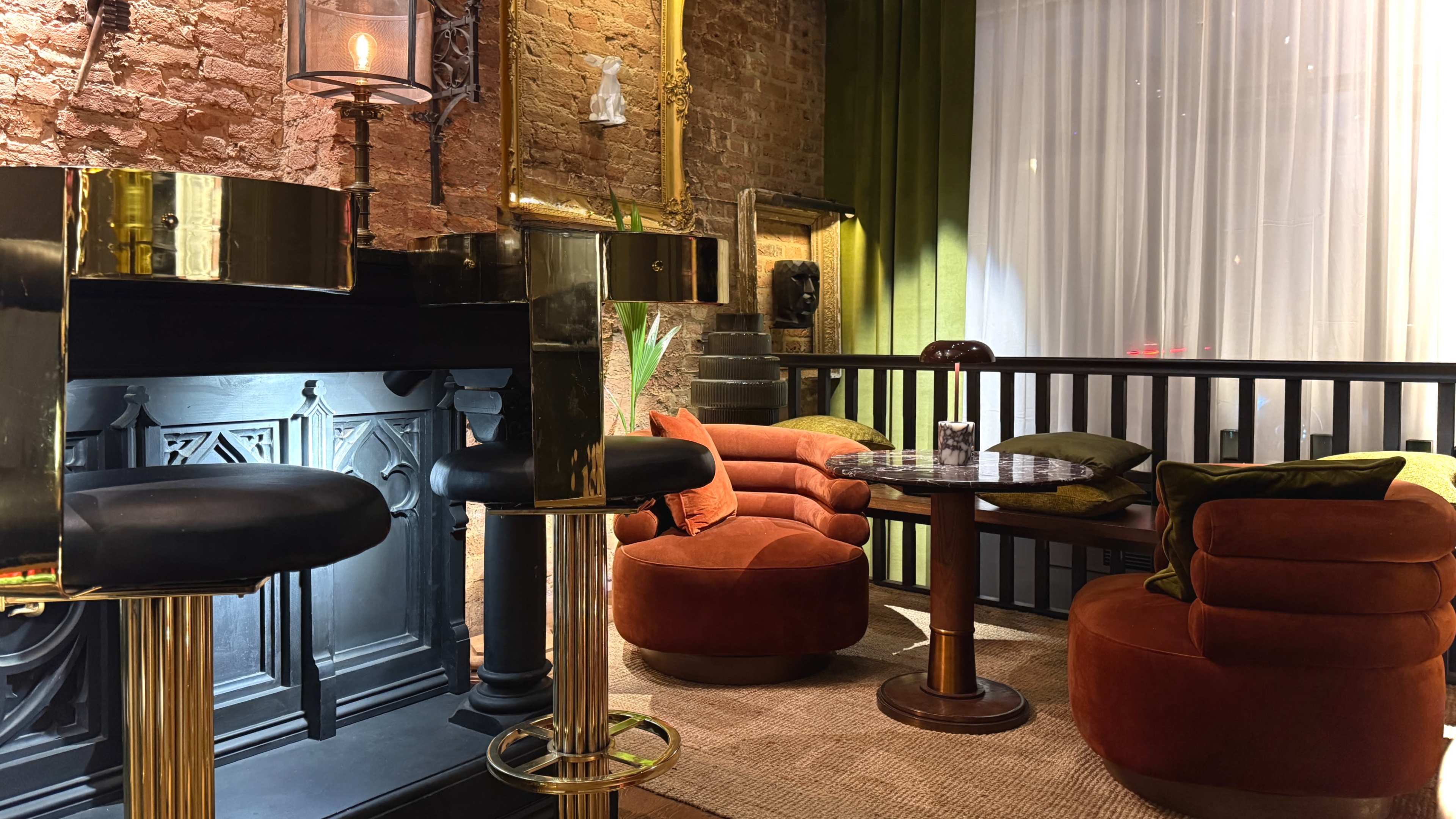 The image shows a stylish interior featuring a bar with gold accents, orange velvet seating, a round wooden table, and a brick wall backdrop.