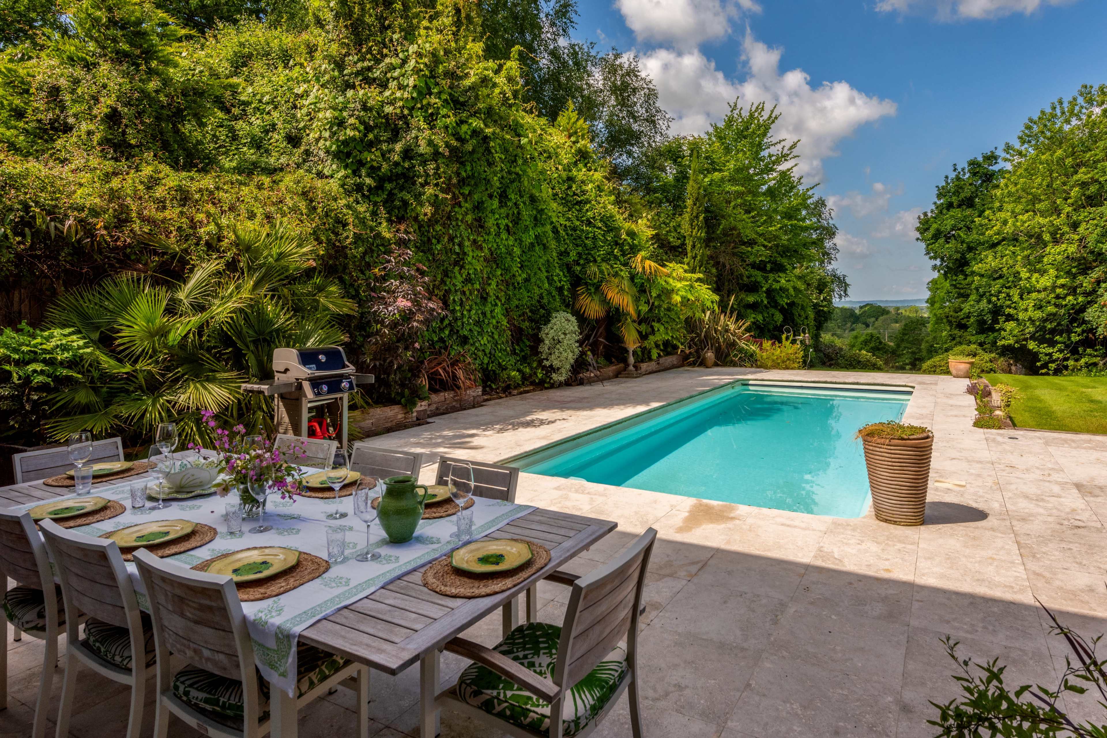 A dining table is set near a swimming pool surrounded by lush greenery and clear skies.