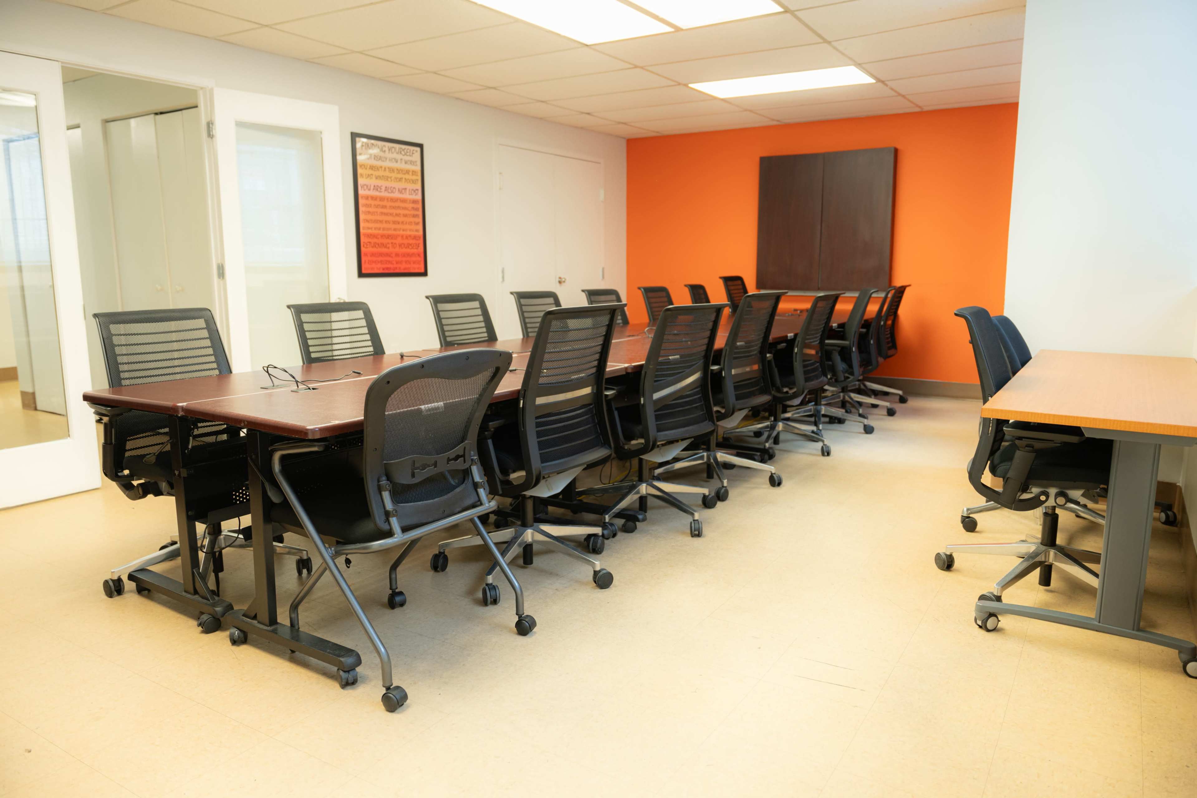 A conference room with a long table surrounded by rolling chairs, featuring a bright orange accent wall.
