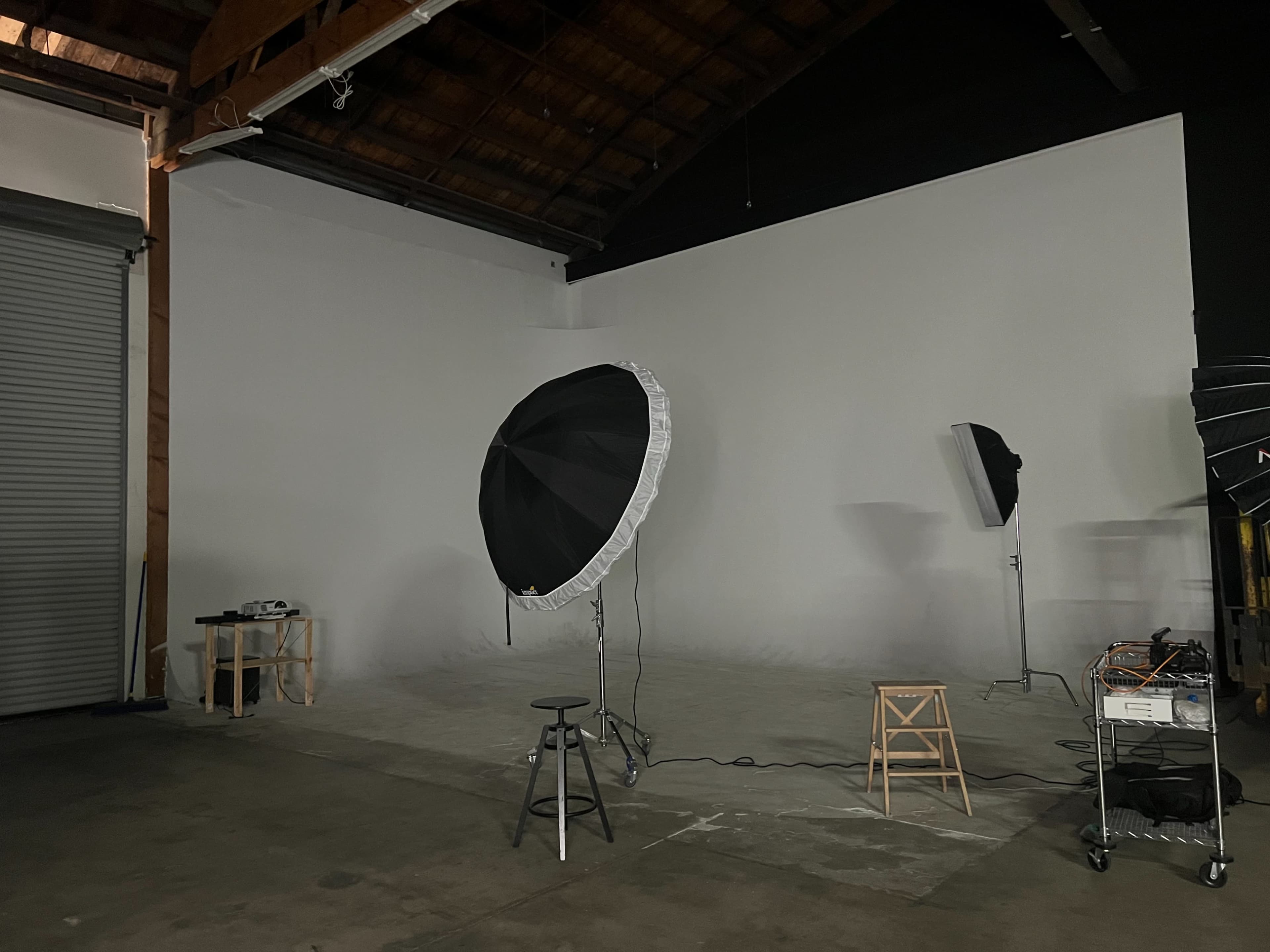 The image shows an empty photography studio with a black umbrella light, a stool, and a backdrop.