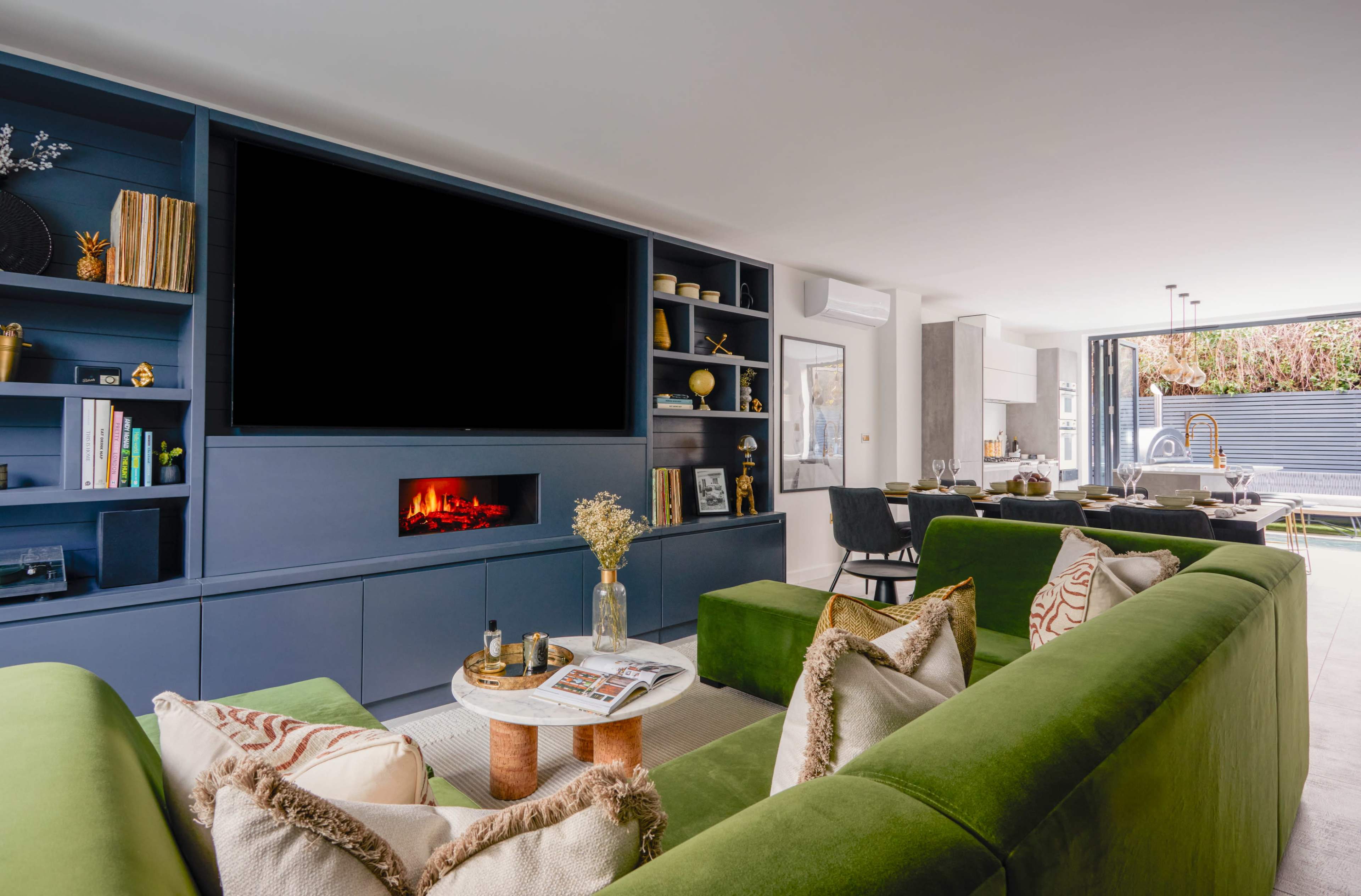 A modern living room features a large dark blue media unit with an empty television space, a coffee table with magazines, and green sofas arranged in a comfortable seating area.