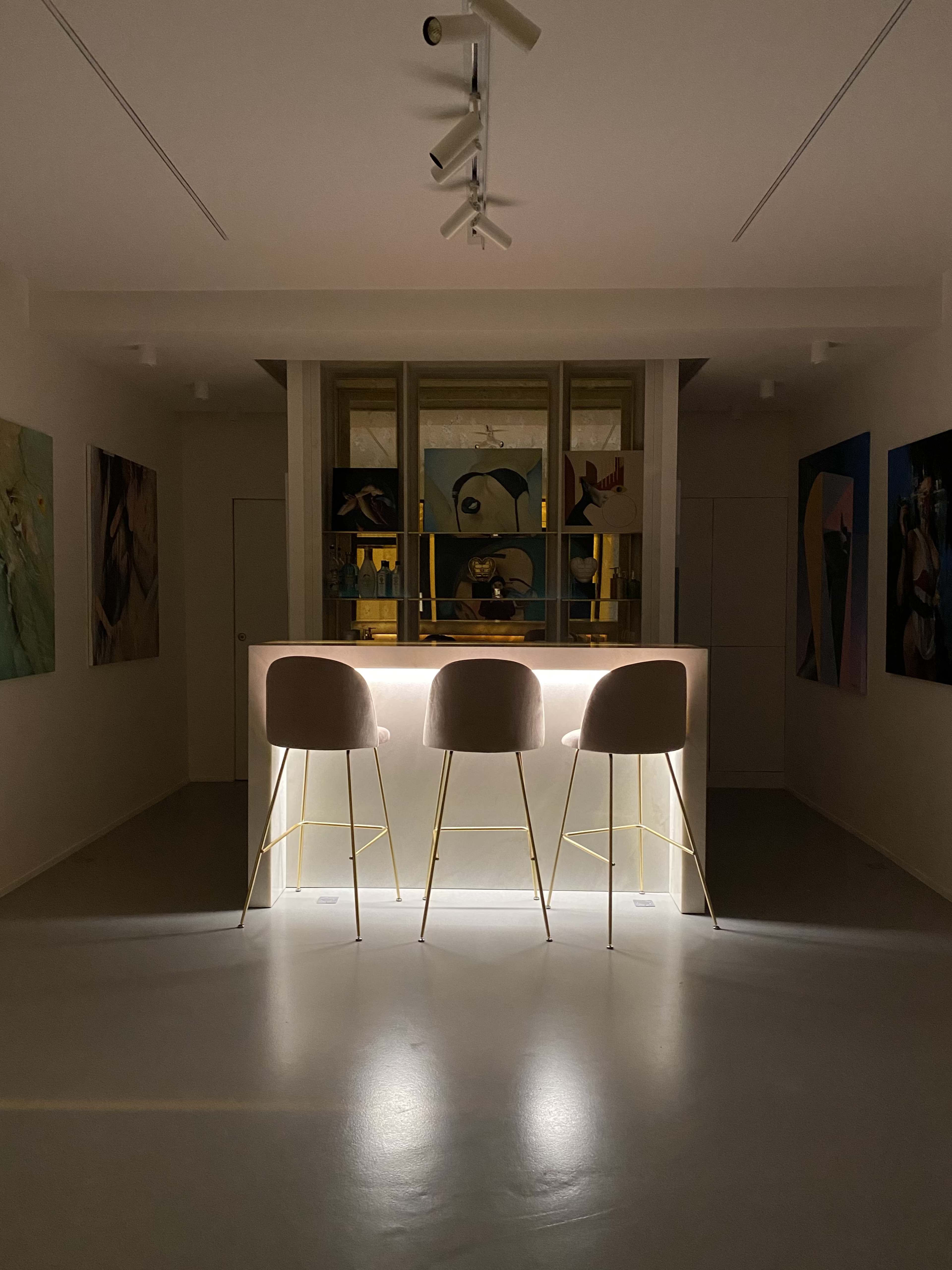 A minimalist bar area with three lighted stools is illuminated, surrounded by various artworks on the walls.