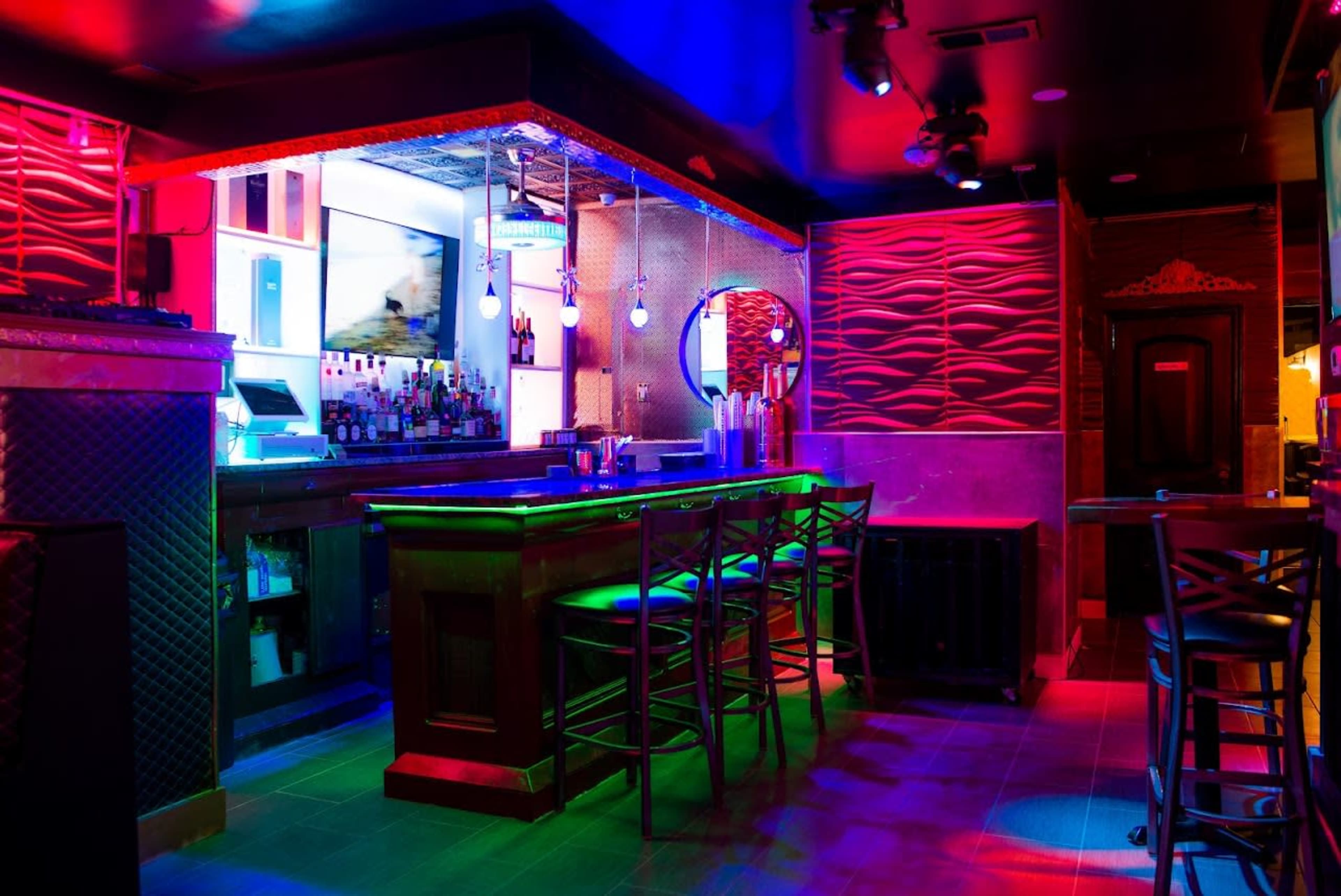 The image shows a dimly lit bar area with colorful neon lighting, a wooden bar counter, high stools, and a variety of liquor bottles on display.