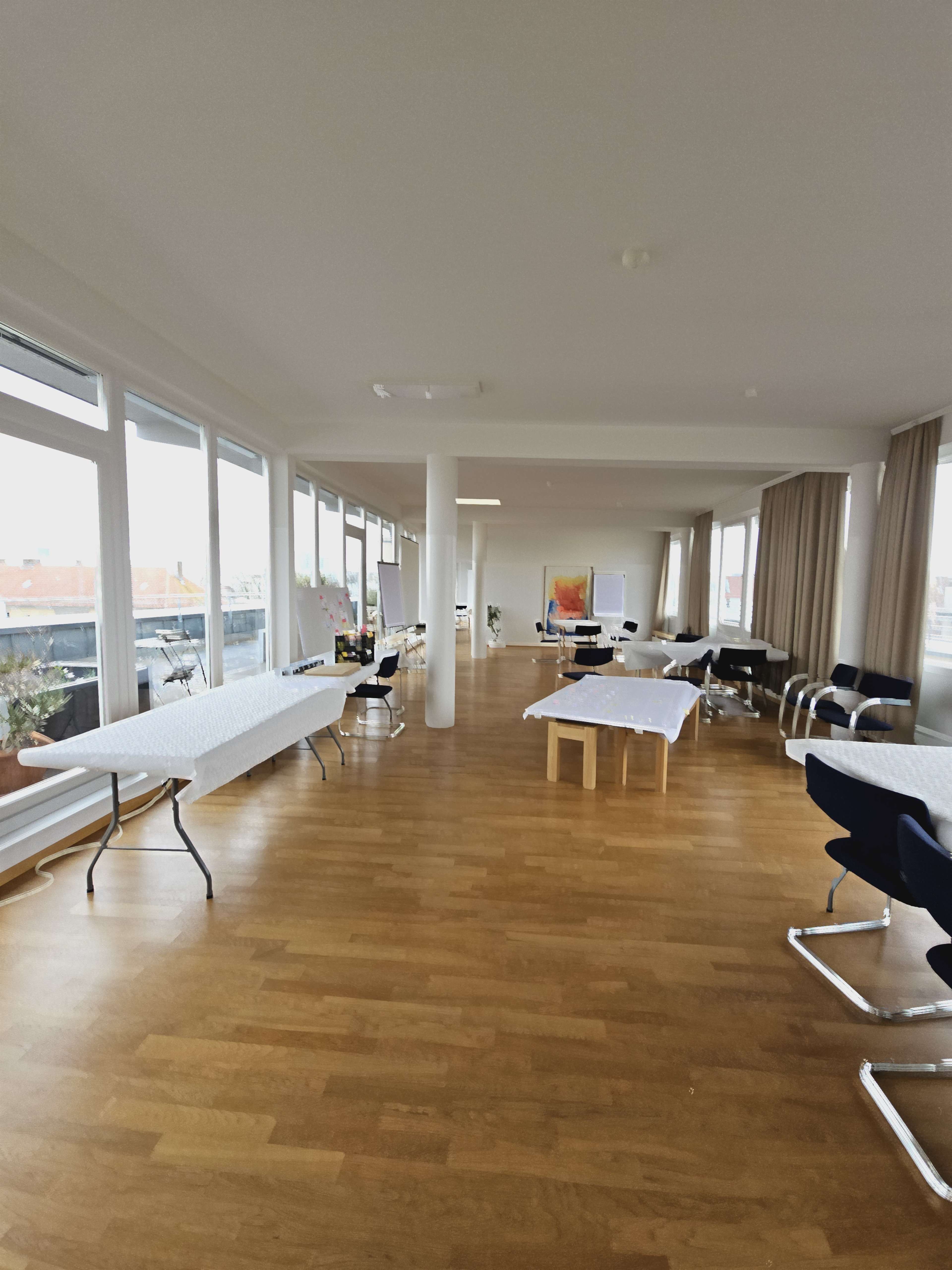 The image shows a spacious room with several tables covered in white cloths and large windows allowing natural light to fill the space.