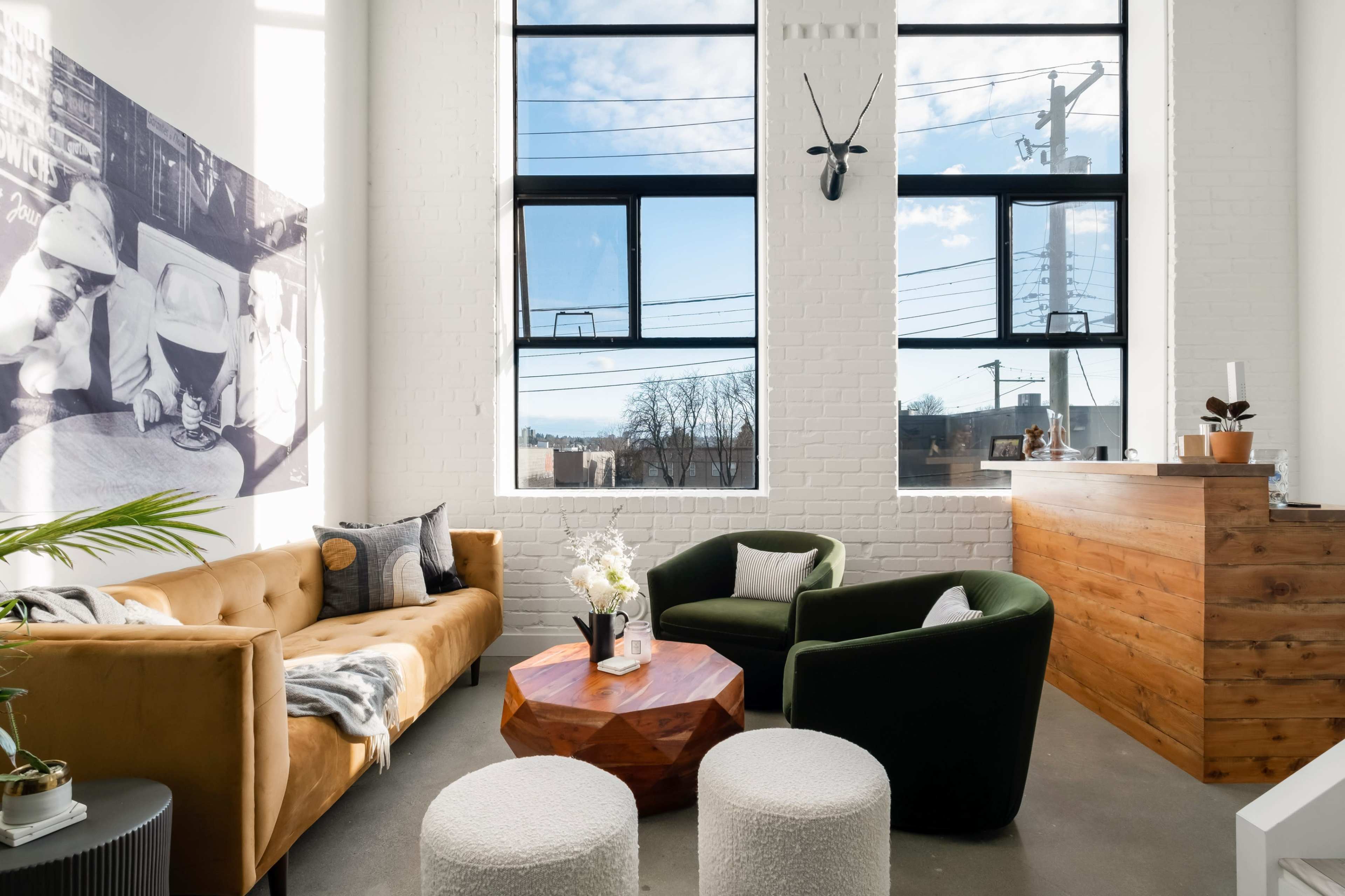 The image shows a modern living room with large windows, a beige sofa, two round white stools, a green armchair, and a wooden reception desk.