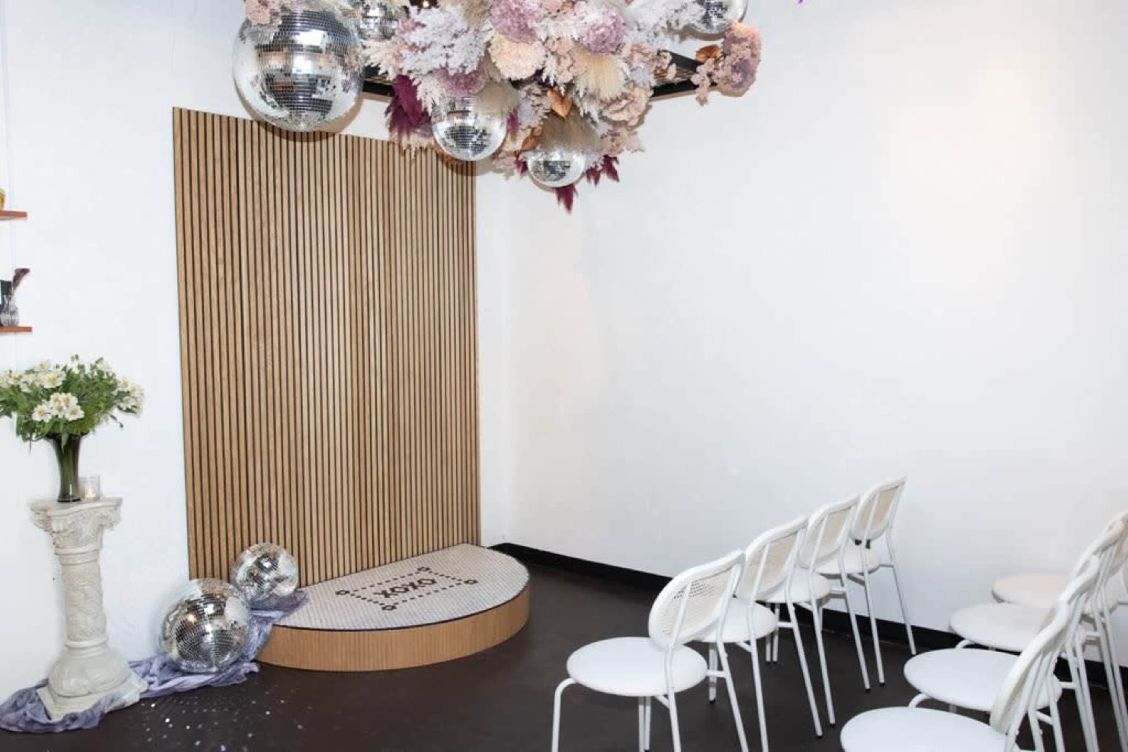 The scene depicts a decorated event space featuring a wooden backdrop with a circular stage, white chairs arranged in rows, and hanging disco balls above.