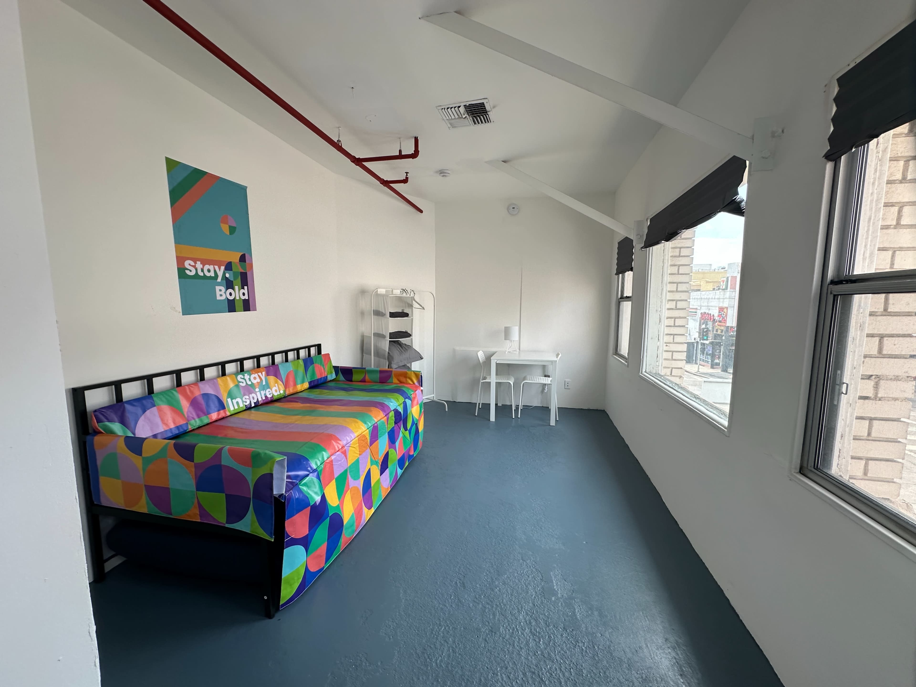 The image shows a bright, minimalistic room featuring a multicolored couch, a small white desk, and large windows that provide natural light.