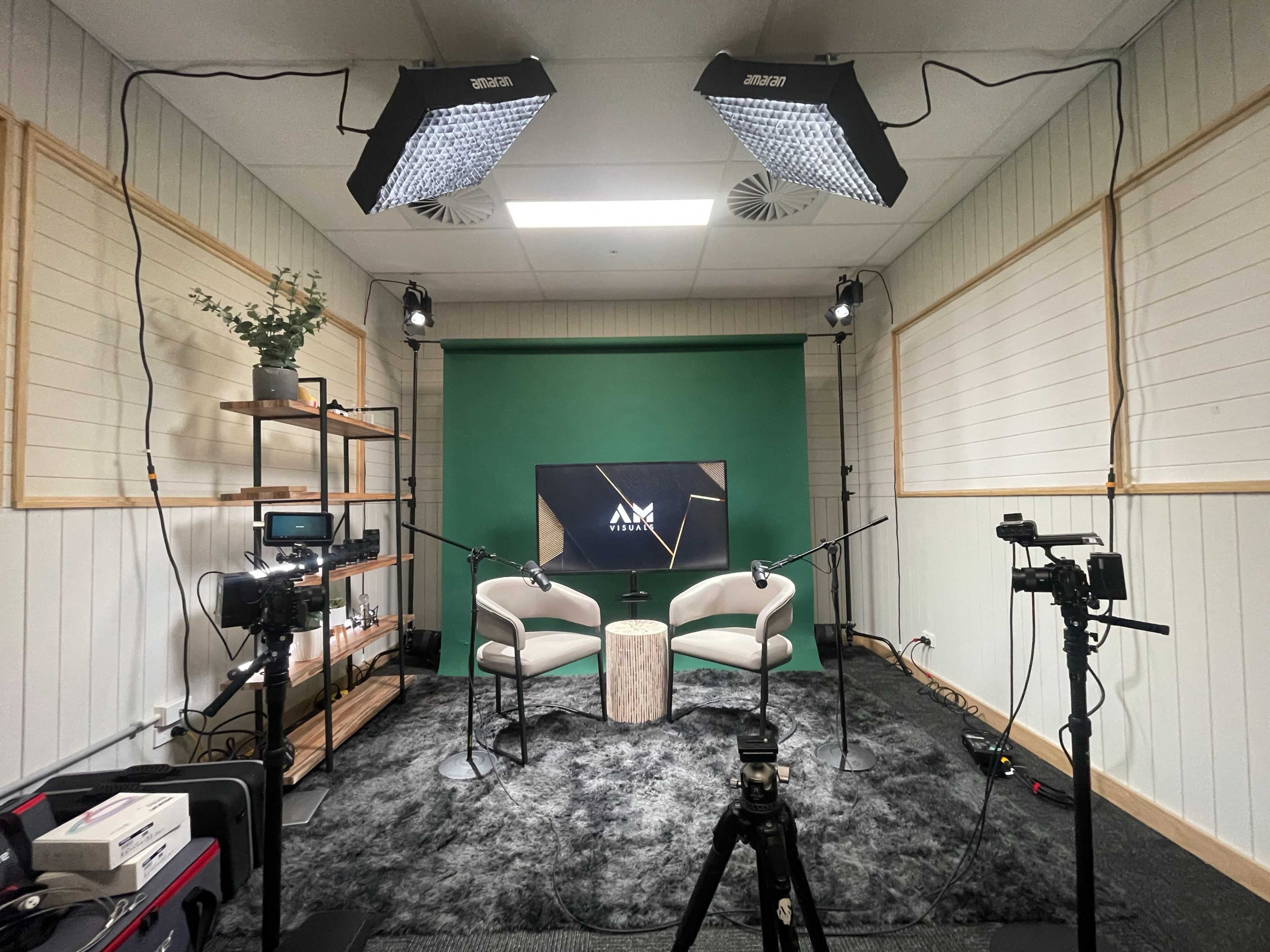 The image shows a podcast studio setup with two white chairs, a green backdrop, and multiple cameras and lighting equipment.