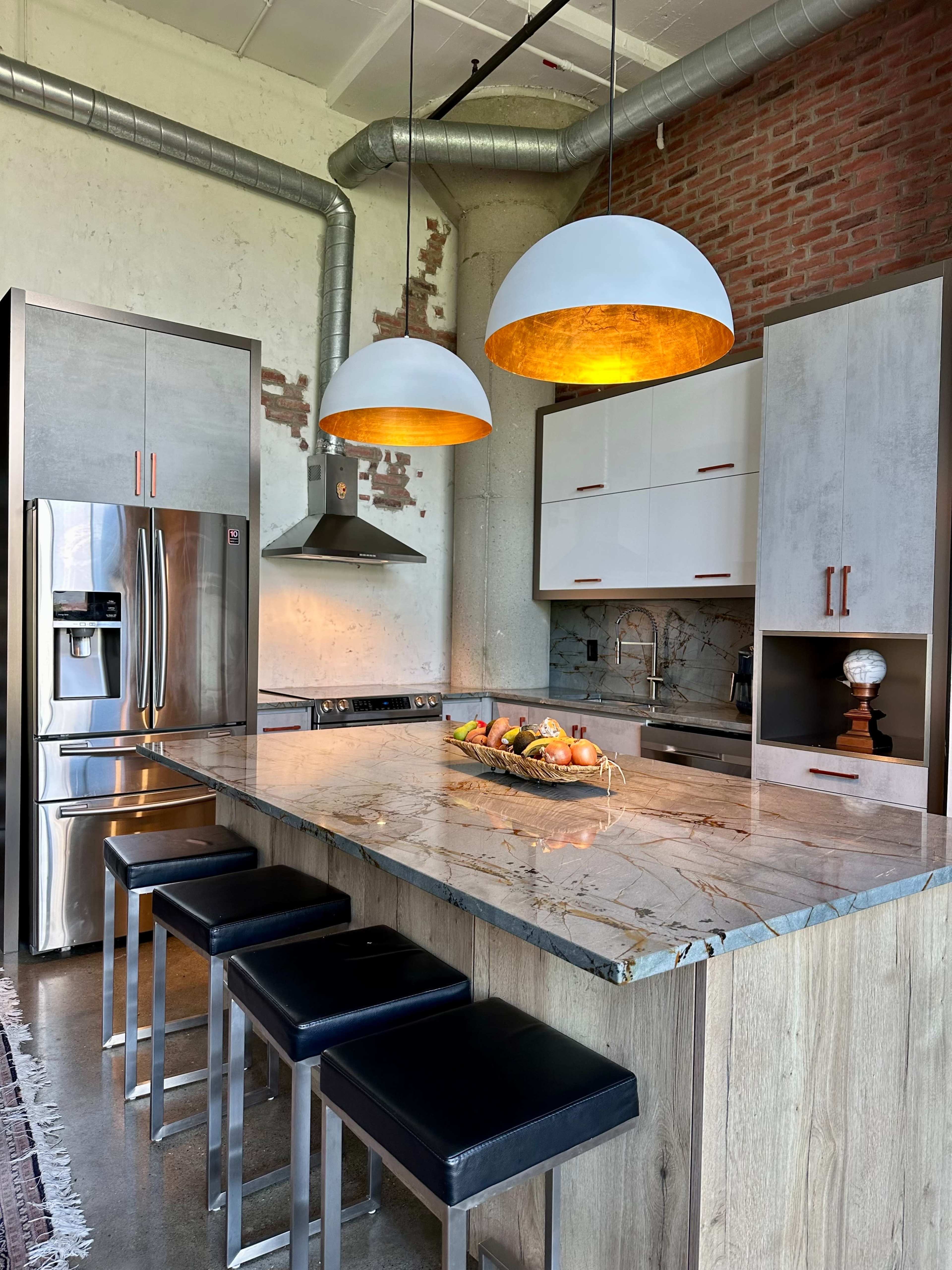 The image shows a modern kitchen with a large island featuring a marble countertop, four black bar stools, stainless steel appliances, and two pendant lights hanging above.