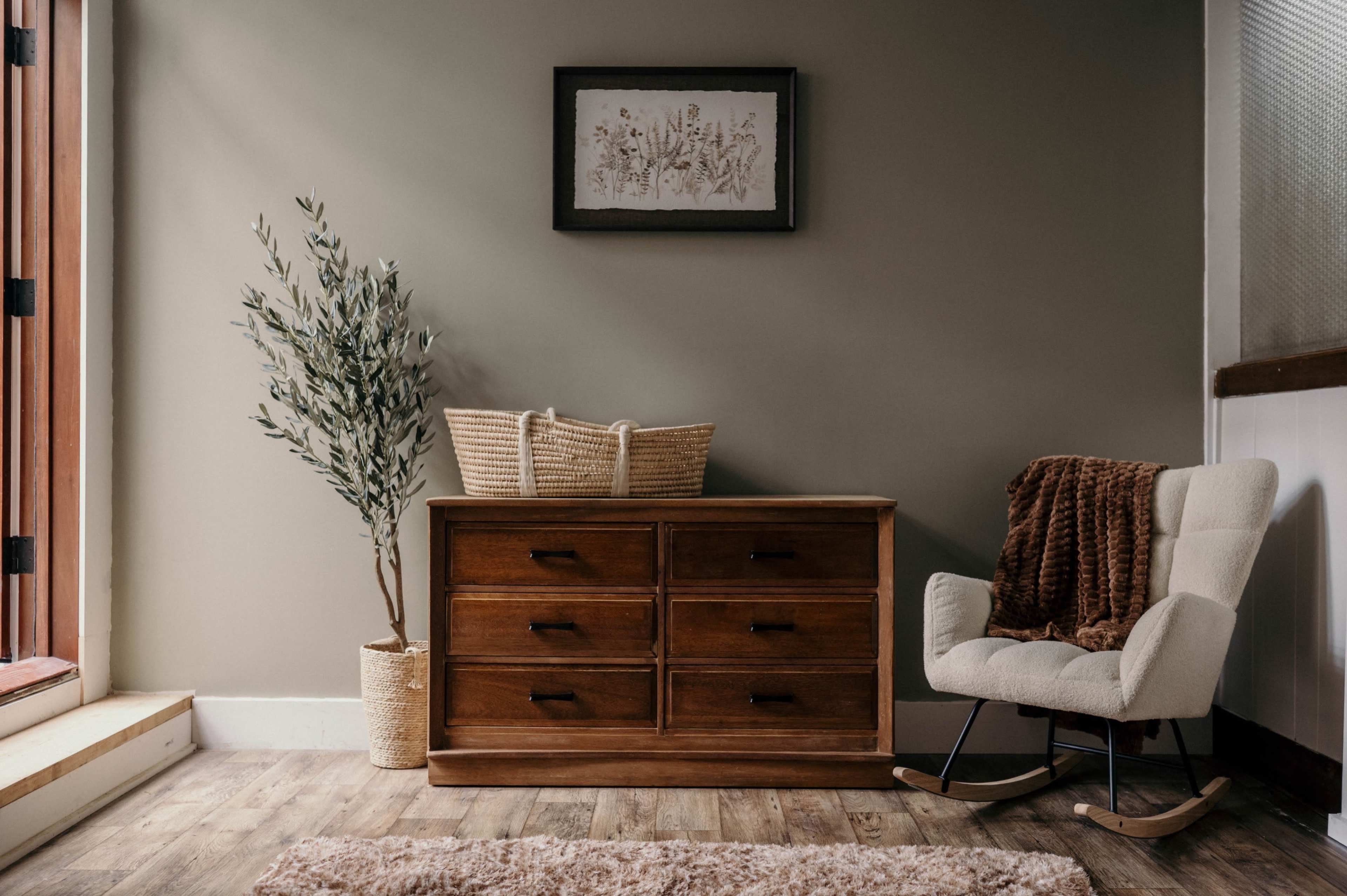 The image shows a wooden dresser with six drawers, accompanied by a decorative plant, a basket, and a rocking chair in a well-lit room.
