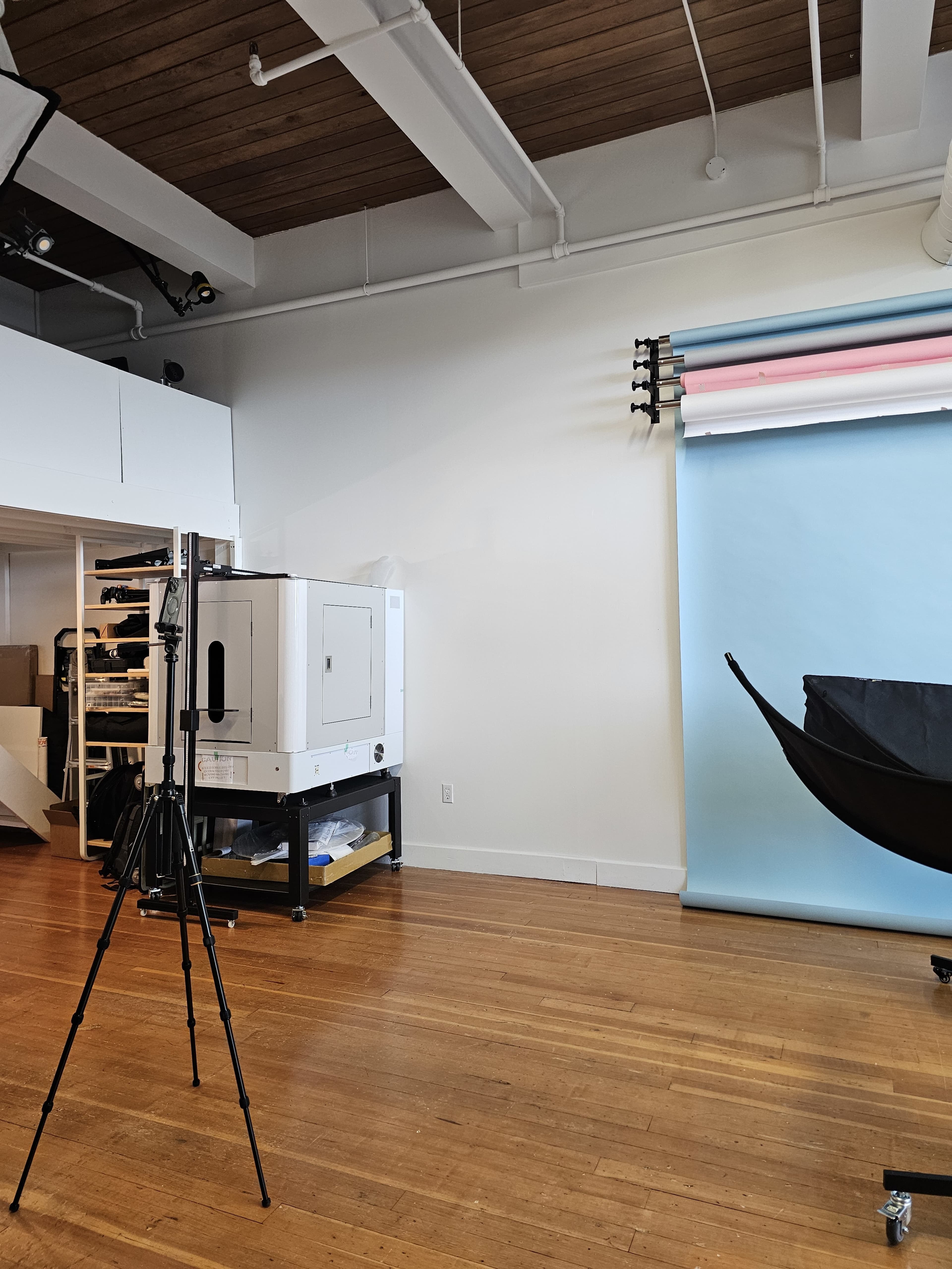 A photography studio with a tripod, a large backdrop, and studio equipment against a light-colored wall and wooden floor.