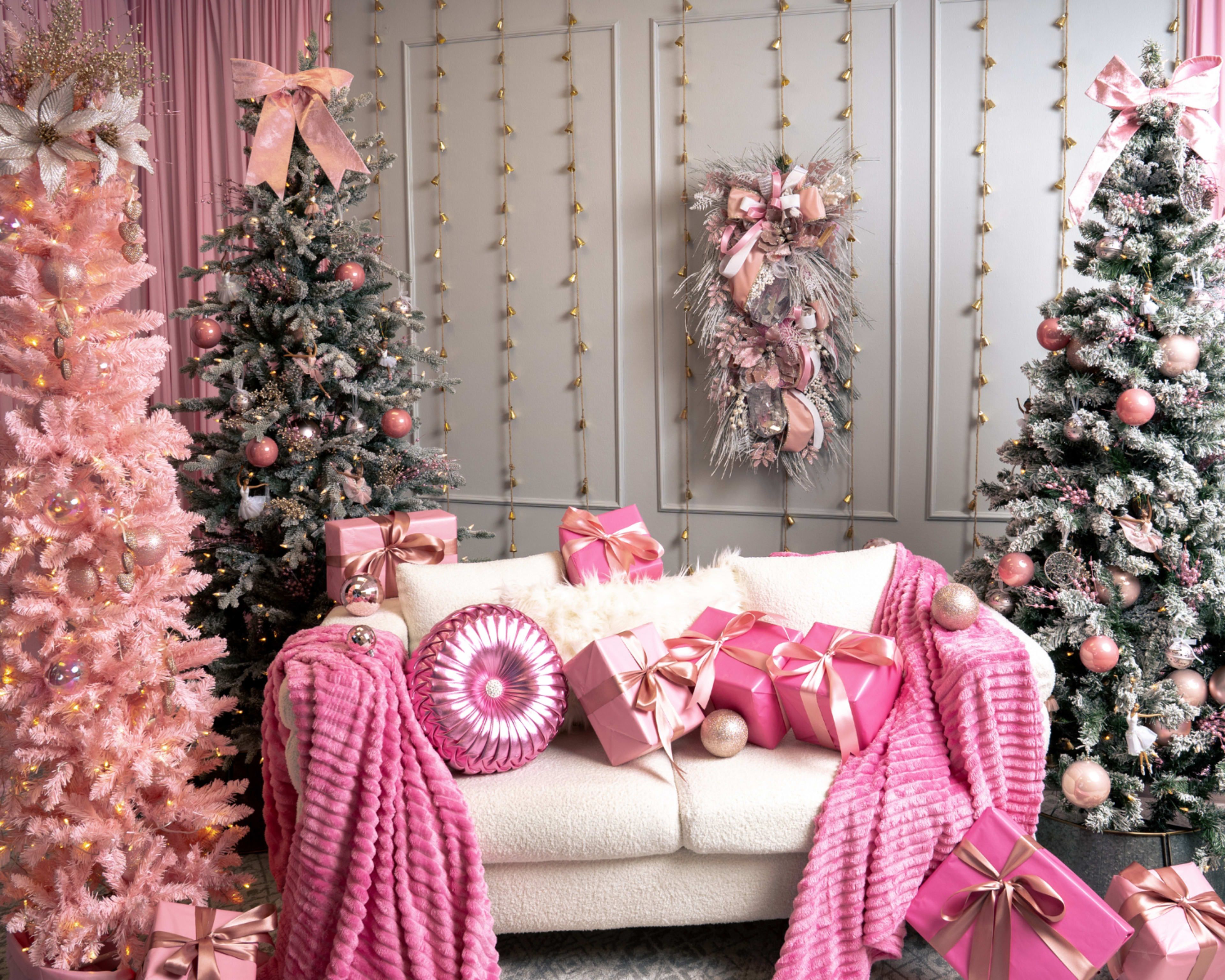 The image shows a festive room decorated for the holidays, featuring pink Christmas trees, a cozy white couch adorned with pink gifts, and a wreath hanging on the wall.