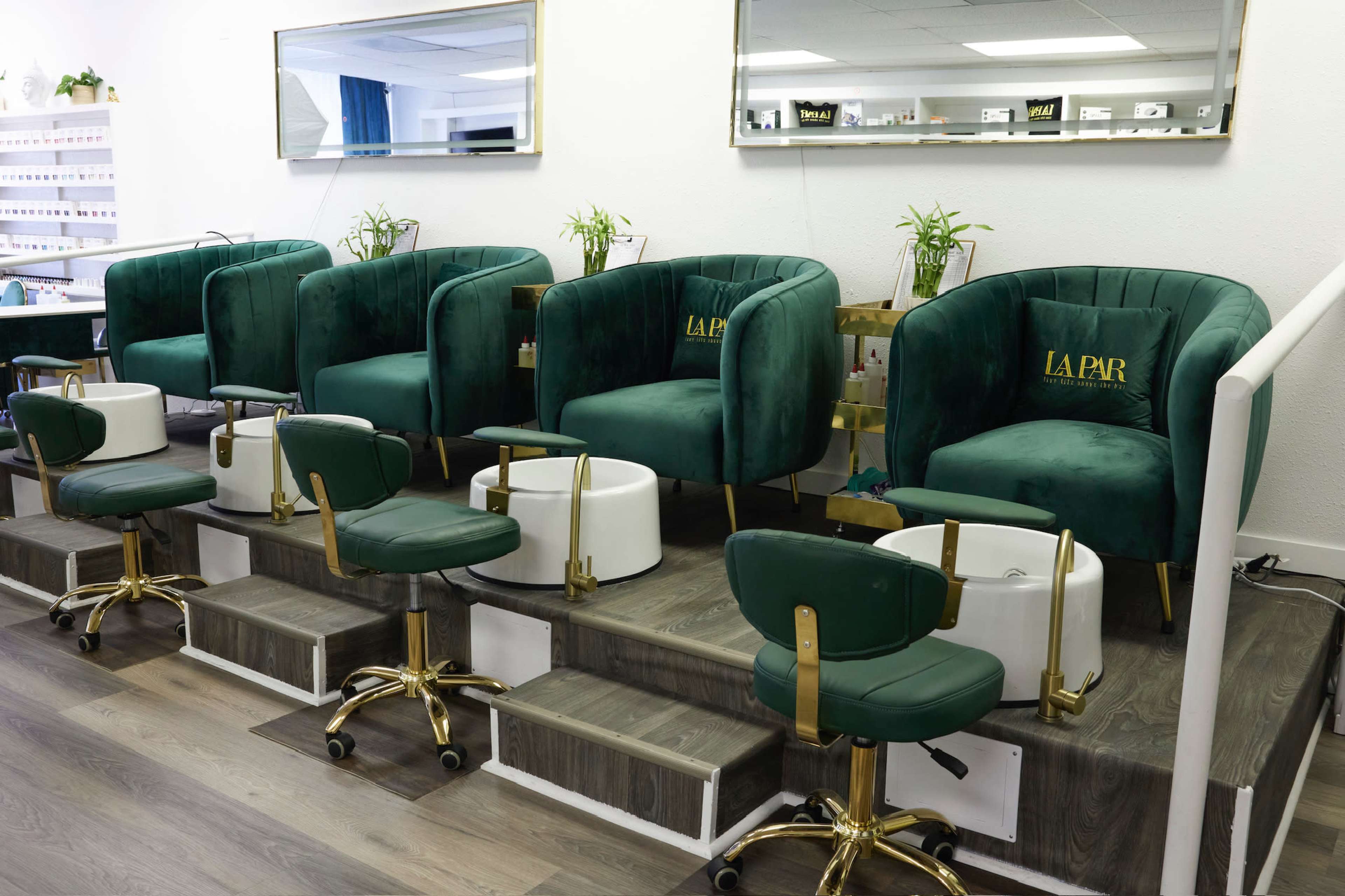 The image shows a nail salon with several green velvet chairs and white pedicure tubs arranged in a line along a stepped platform.