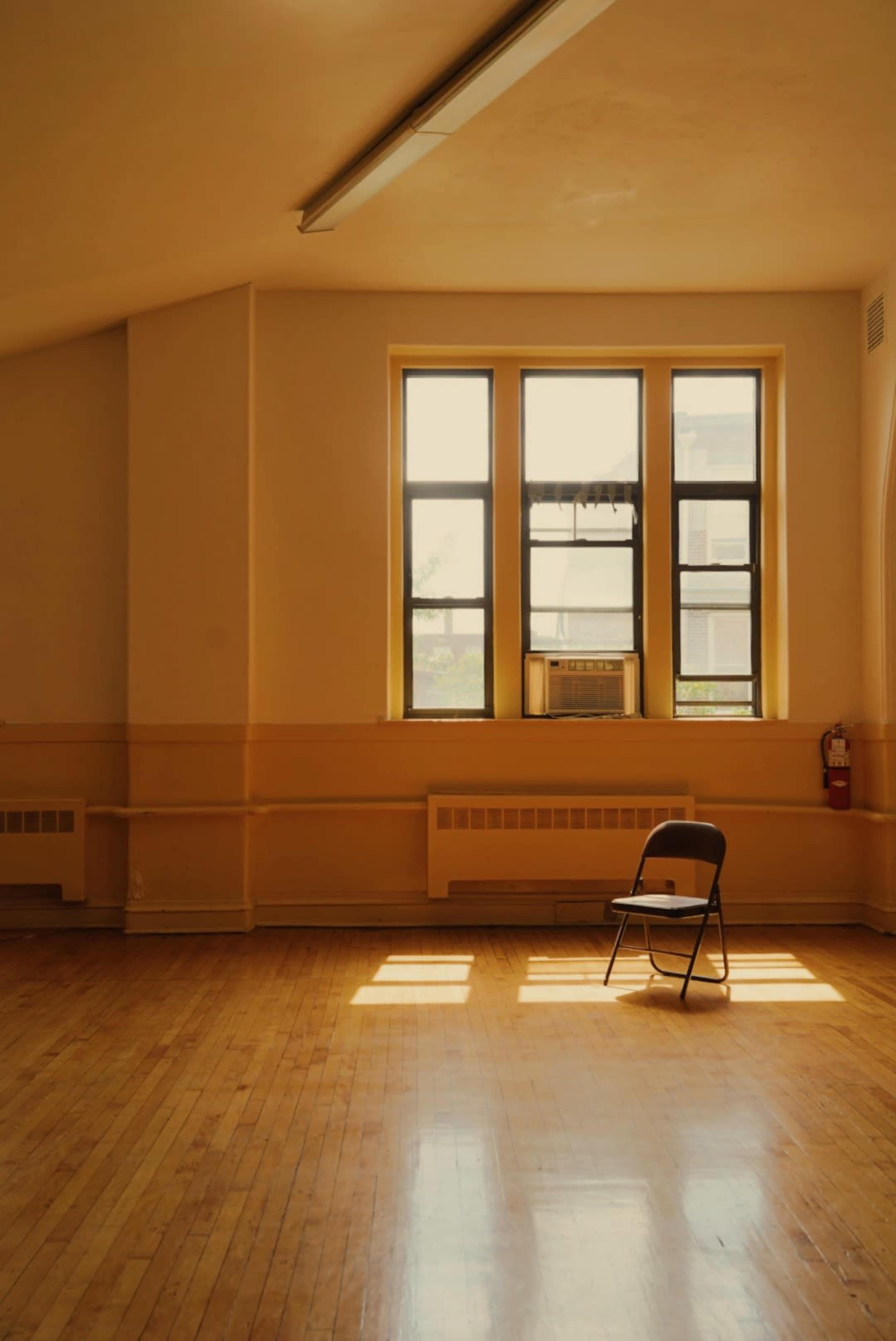 A single black chair is positioned on a wooden floor in a sunlit, empty room with large windows.