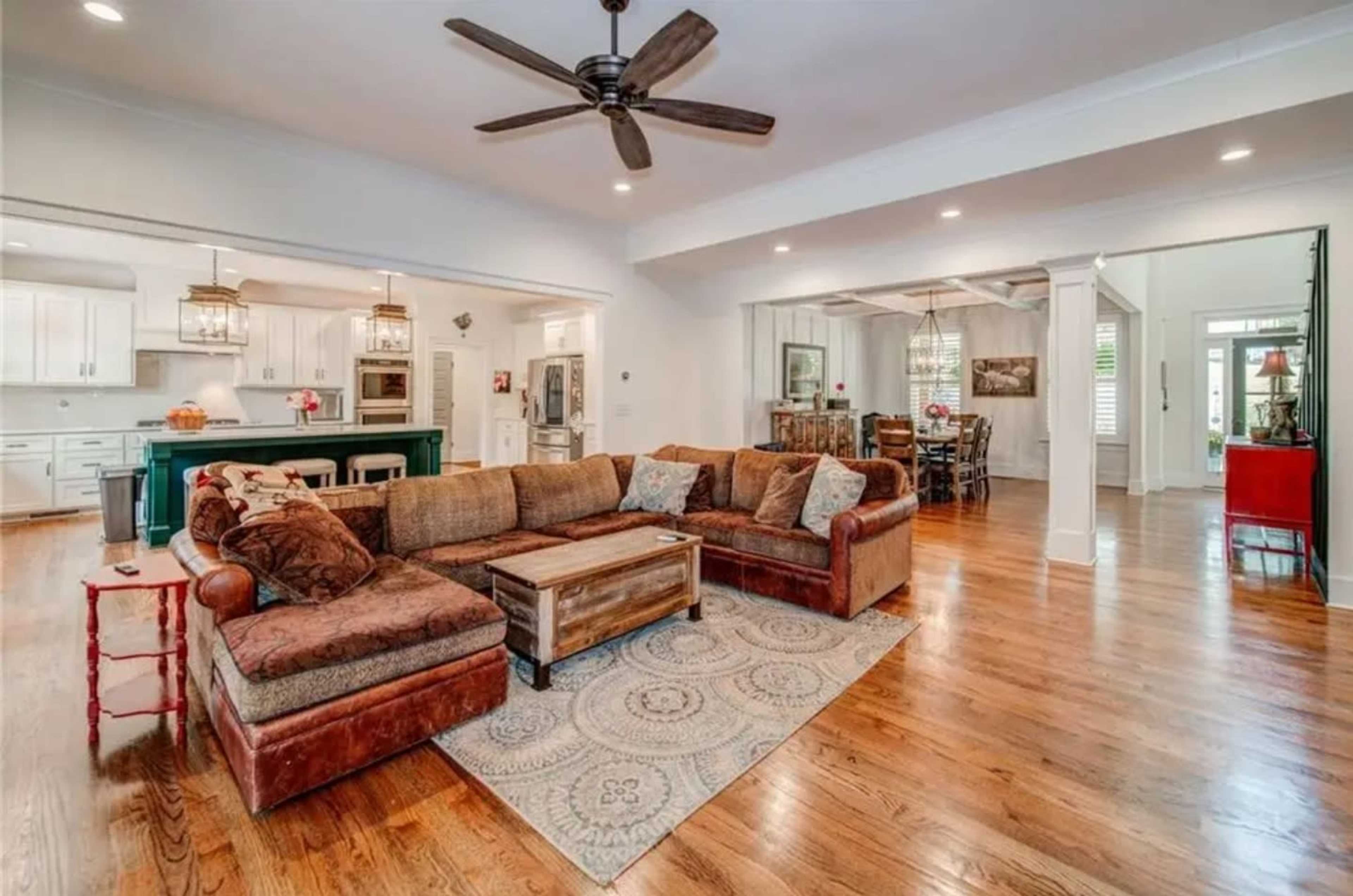 The image shows a spacious living area featuring a large sectional couch, a coffee table, and an open layout that connects to a kitchen and dining area.