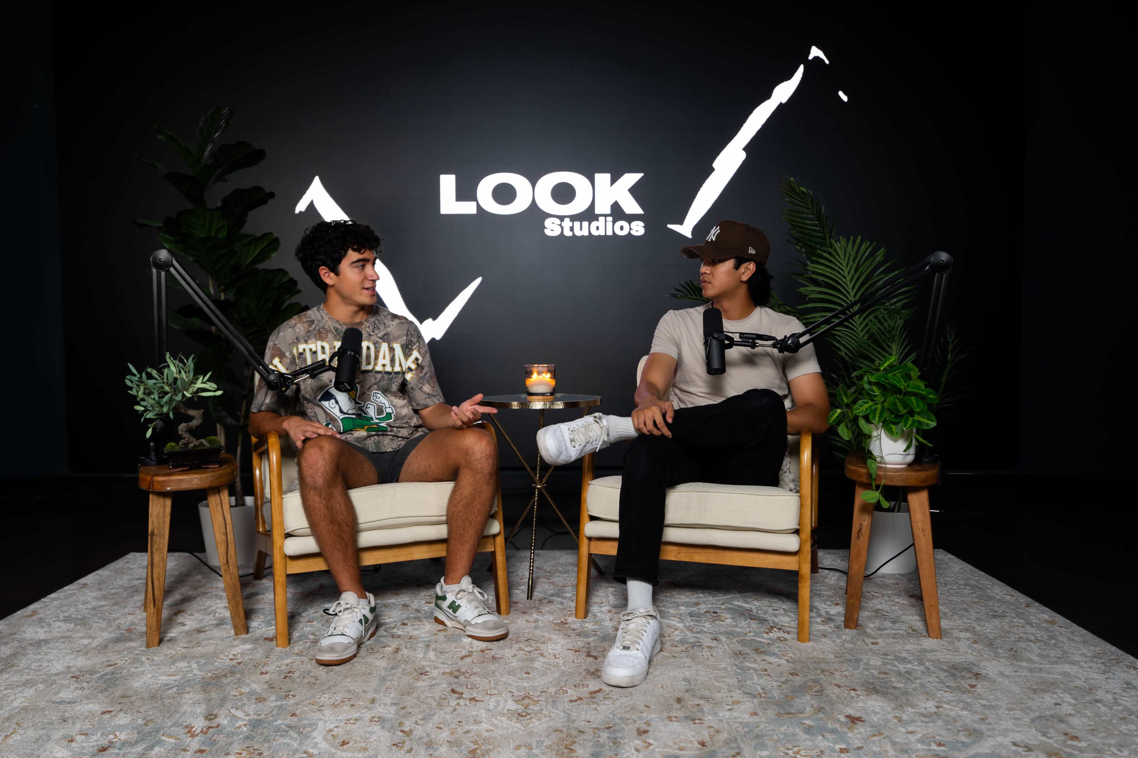 Two young men are seated in armchairs in a modern studio setting, with microphones positioned in front of them and a large "LOOK Studios" logo on the wall behind.