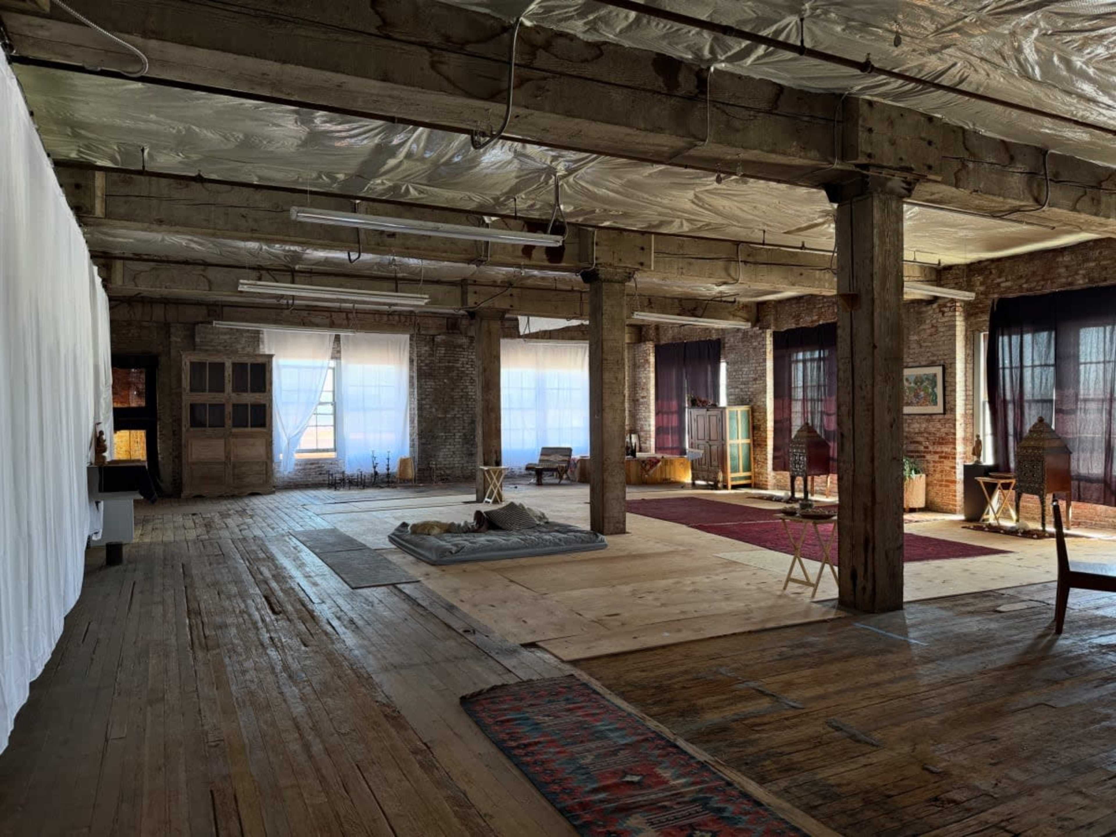 The image shows an expansive, industrial-style room with wooden floors, exposed beams, and large windows allowing natural light to enter.