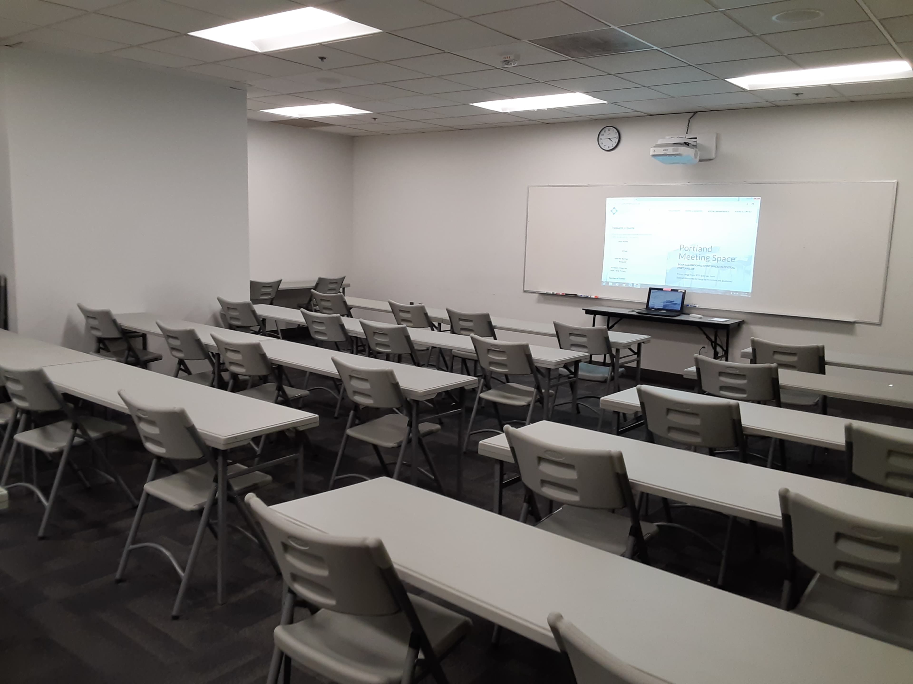 A classroom with multiple rows of folding tables and chairs, a projector setup, and a whiteboard at the front.