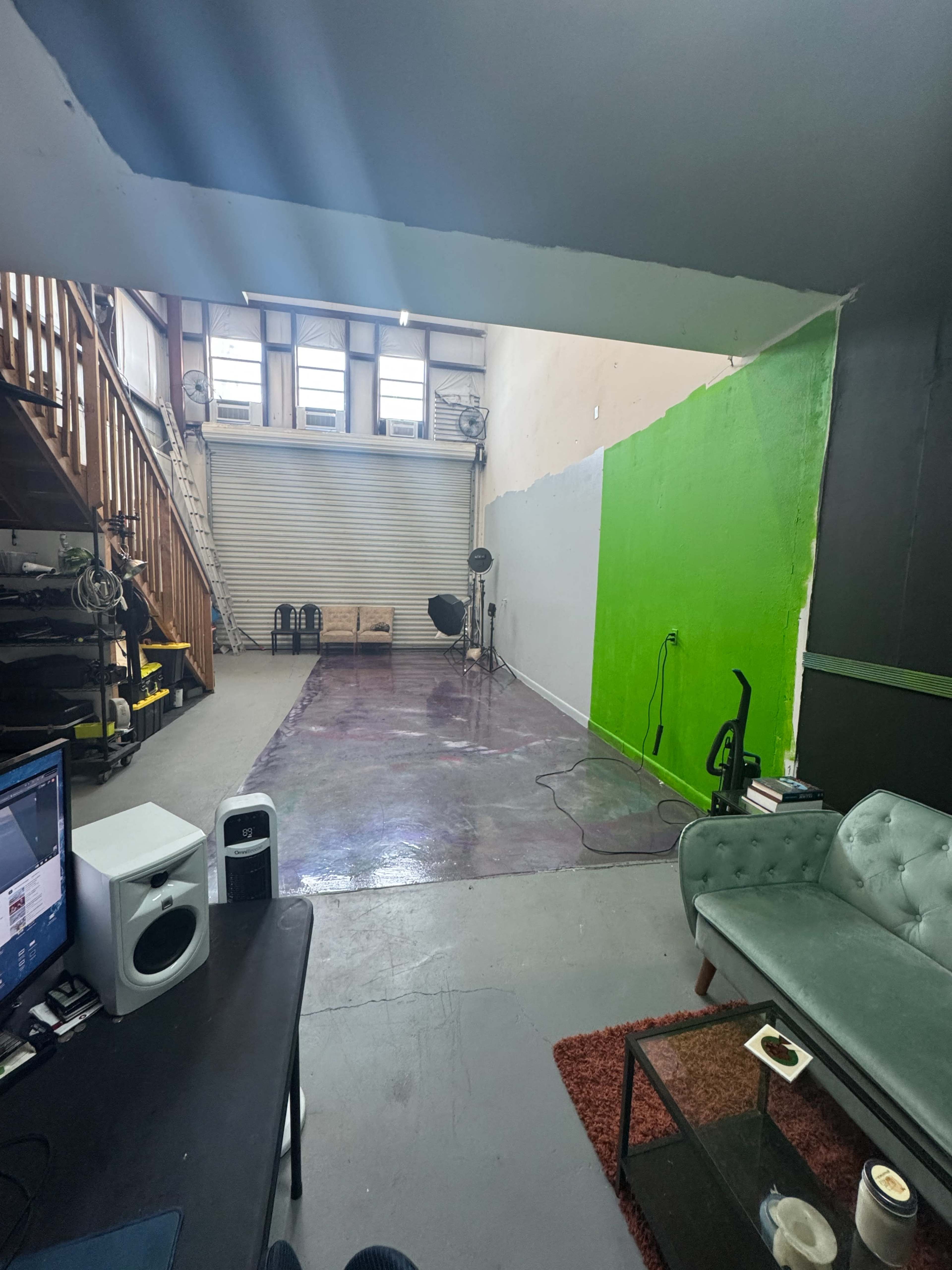 The image shows a spacious studio with a green wall, a staircase on the left, a few lighting equipment pieces, and a teal couch in the foreground.