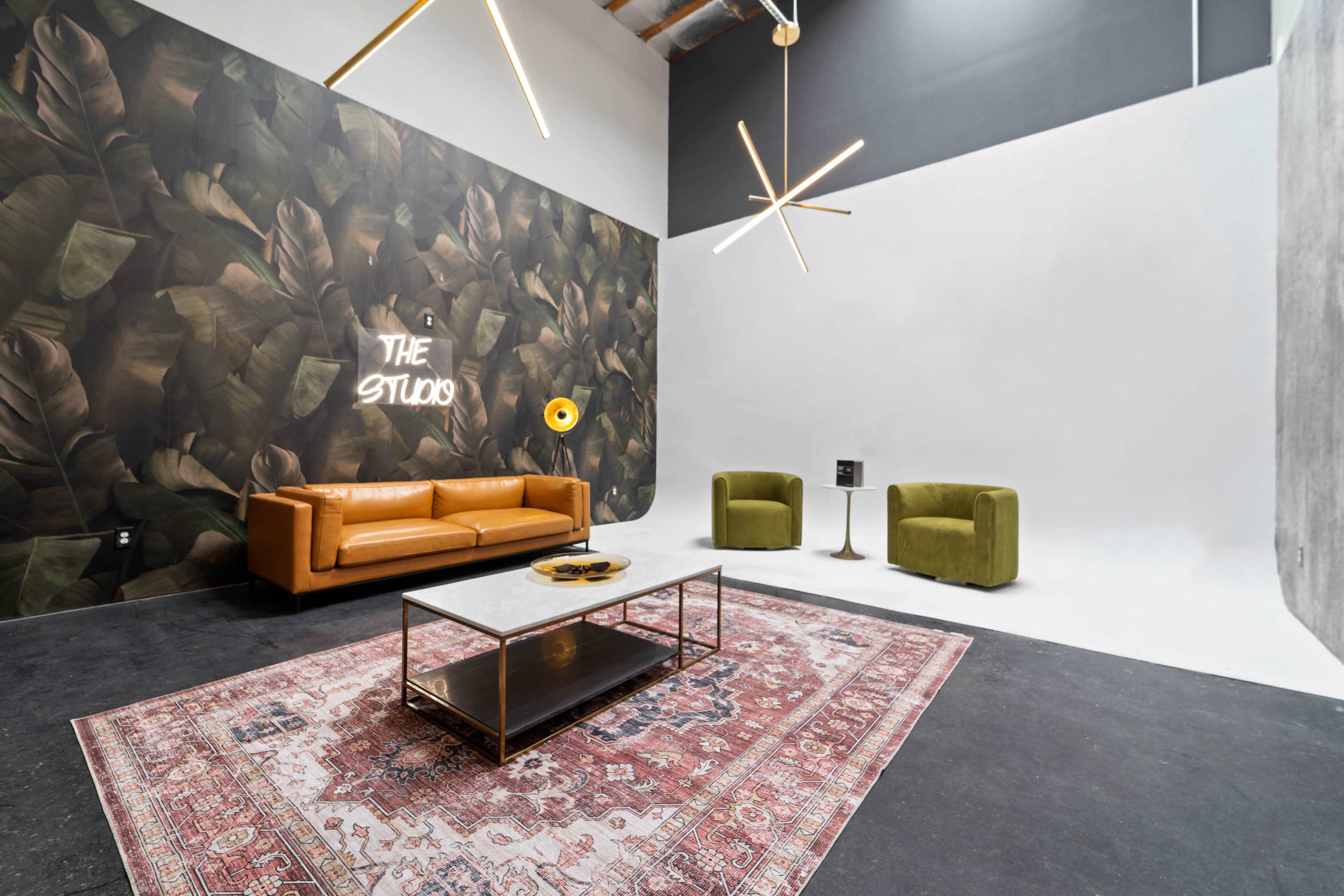 A modern studio space featuring a brown leather sofa, two green chairs, a coffee table with decorative items, and a large mural of tropical leaves on one wall.