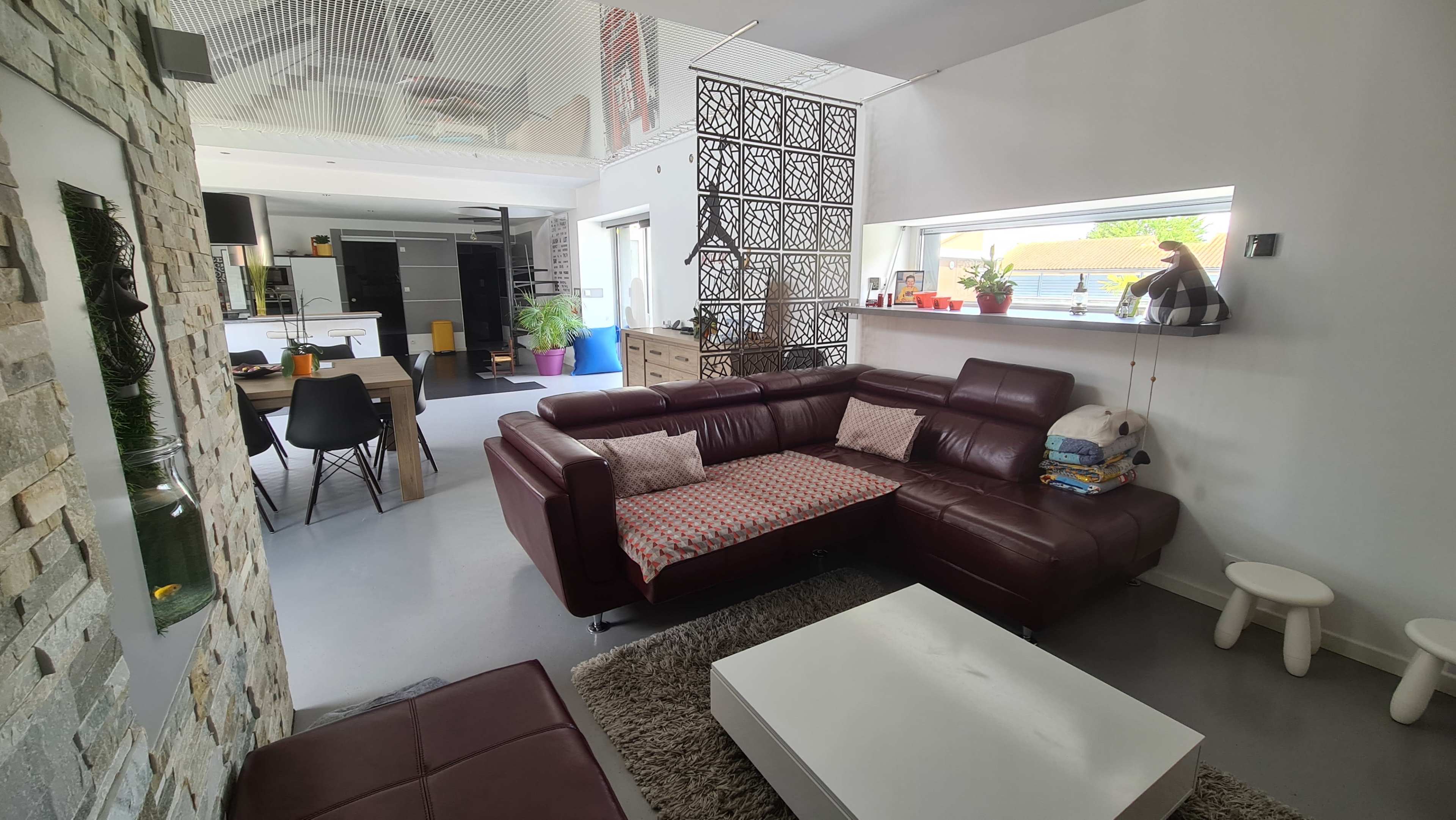 The image shows a modern living space with a brown leather sectional sofa, a white coffee table, and a dining area in the background featuring a wooden table and black chairs.