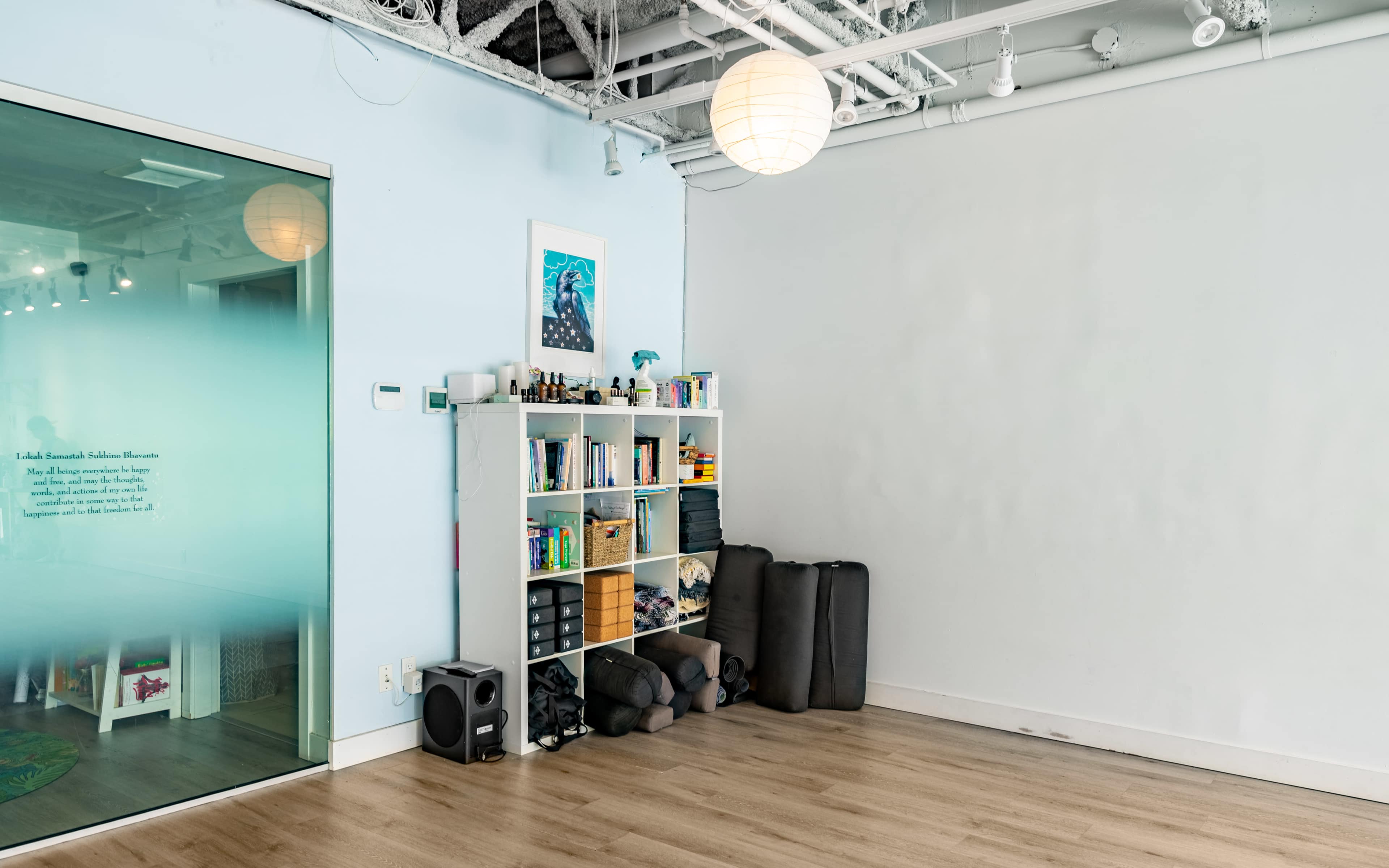 A fitness studio interior features a shelving unit filled with books and equipment next to a stack of exercise mats against a blank wall.