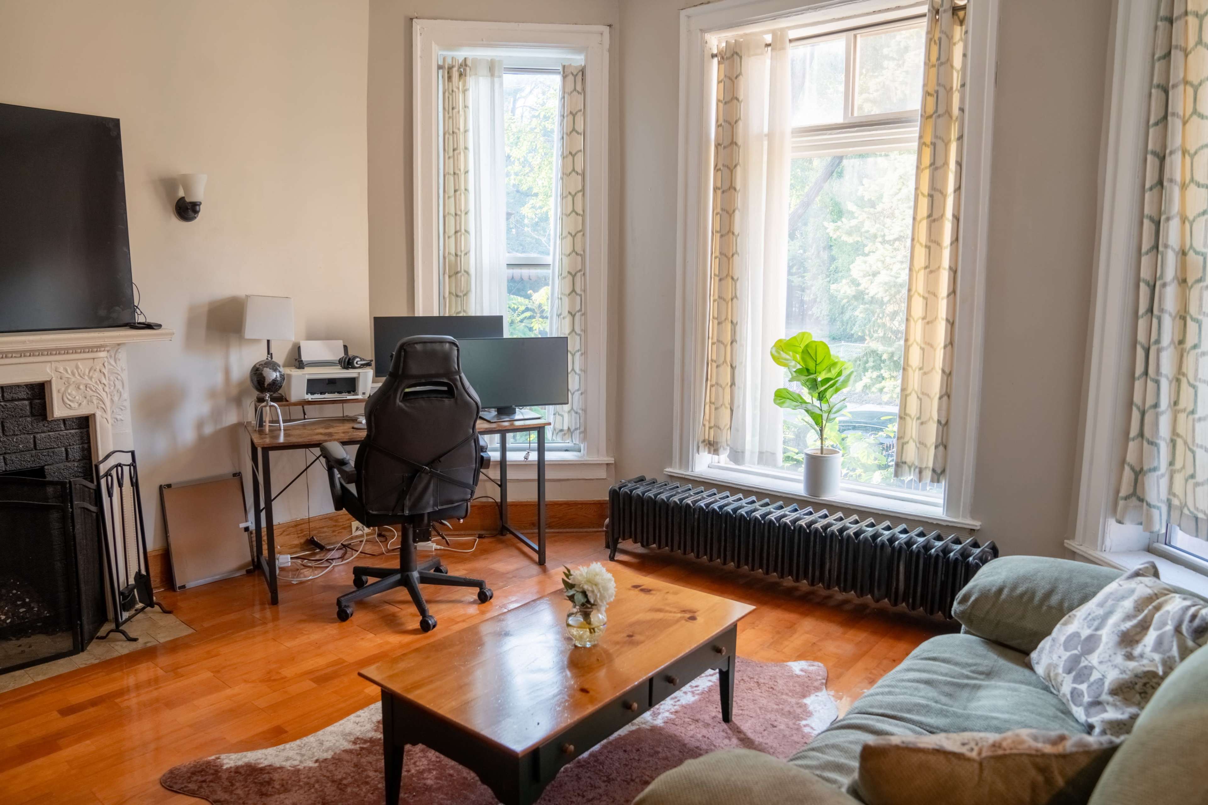 The image shows a cozy living room featuring a green sofa, a wooden coffee table, a desk with a computer setup, and large windows with curtains letting in natural light.