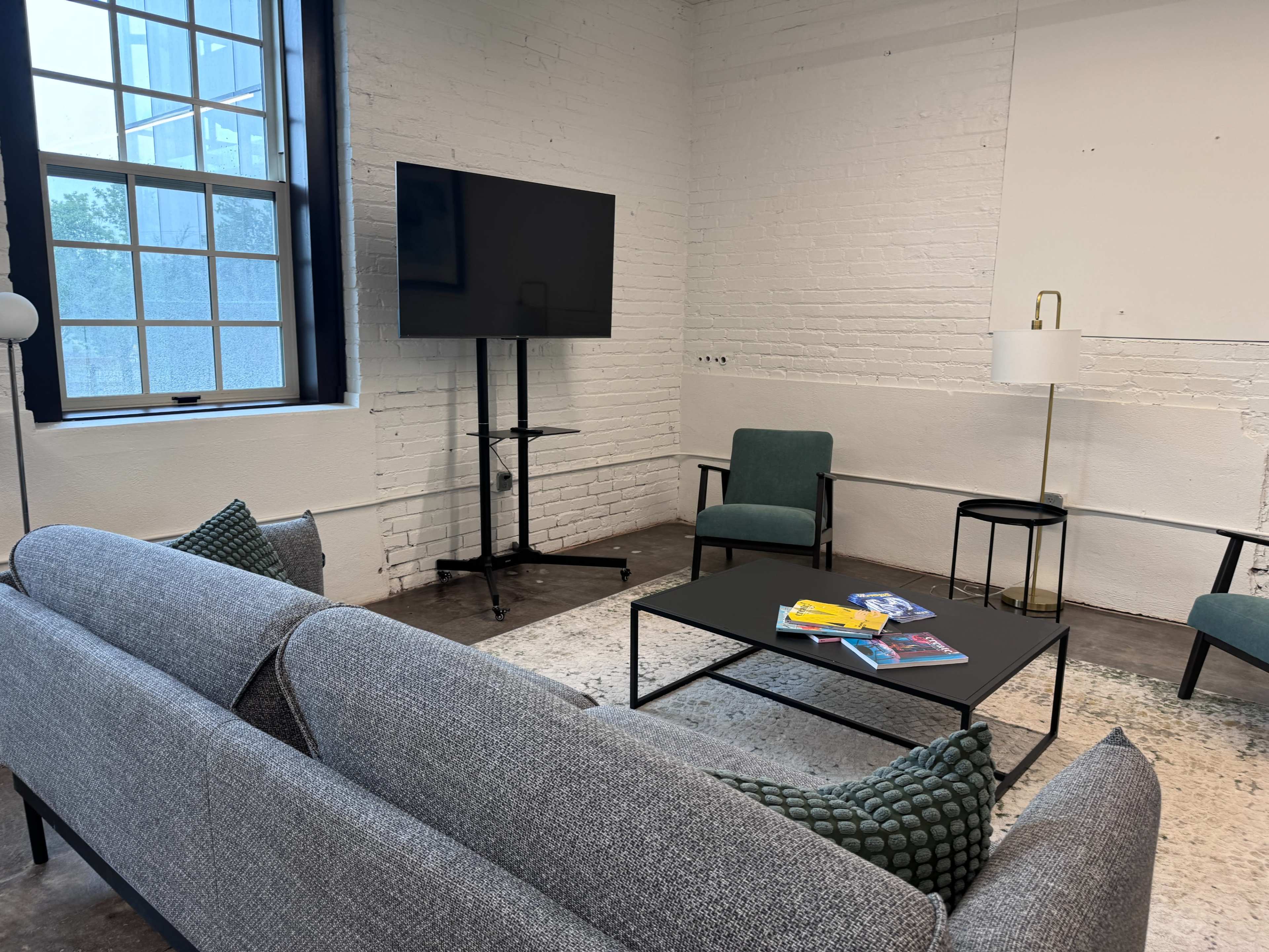 The image shows a modern, minimalist lounge area with a gray sofa, chairs, a coffee table, and a television on a stand, all set against a white brick wall.
